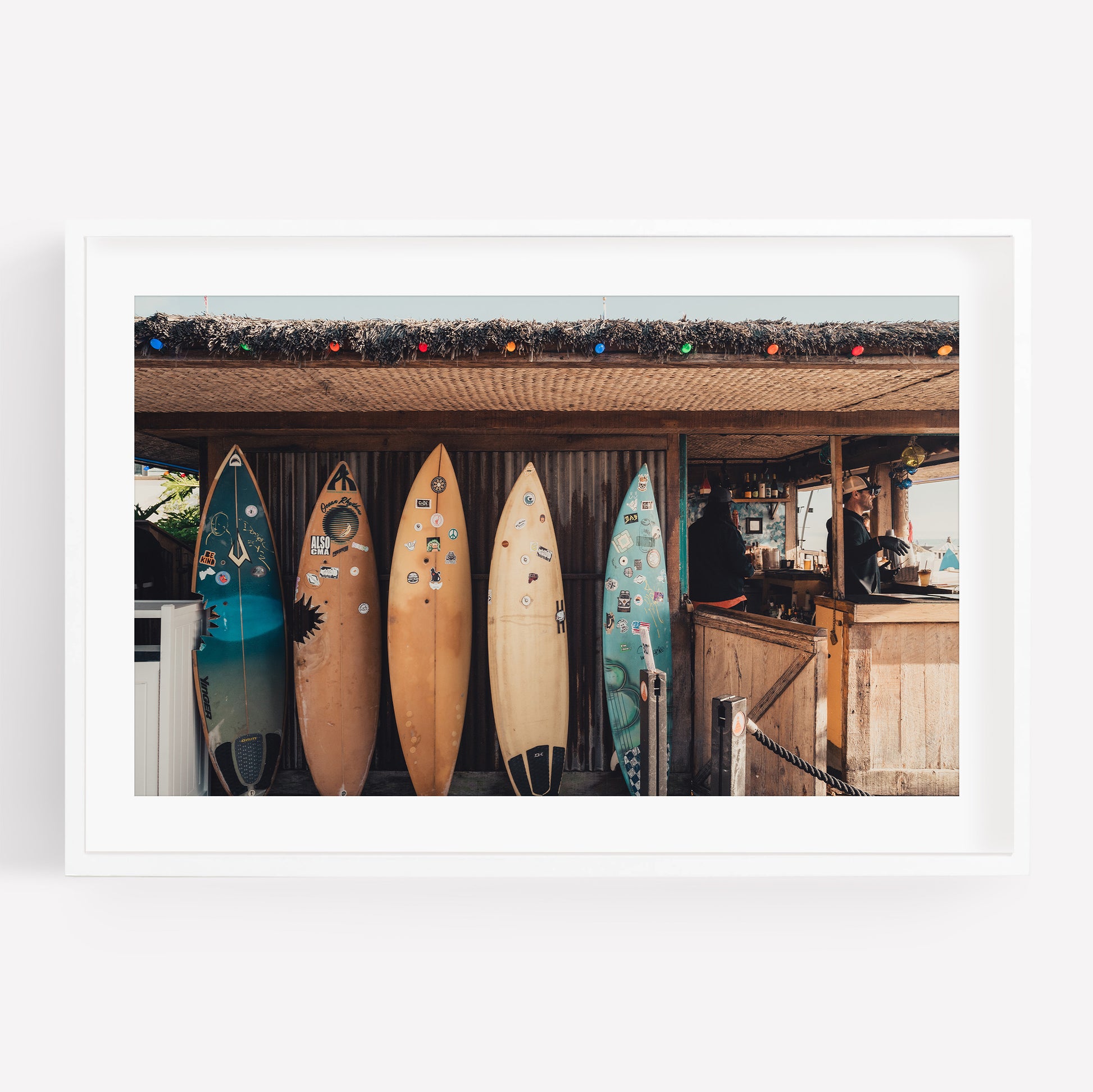 Four surfboards lean against the Surfboard Cafe, adorned with colorful string lights and a person behind the counter.