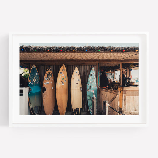 Four surfboards lean against the Surfboard Cafe, adorned with colorful string lights and a person behind the counter.