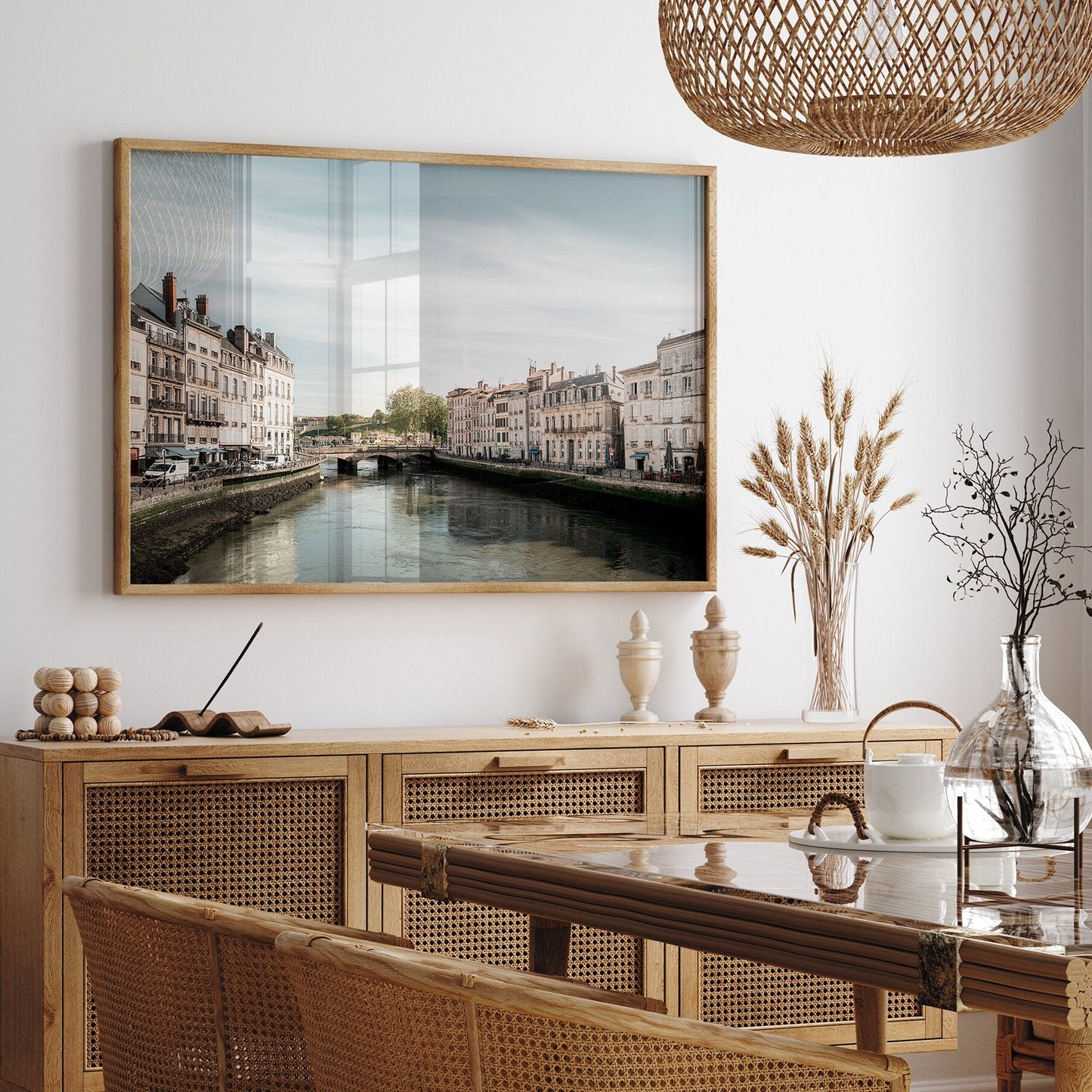 The Adour River photograph is framed on a wall above a rattan sideboard, with vases and a wicker chandelier.