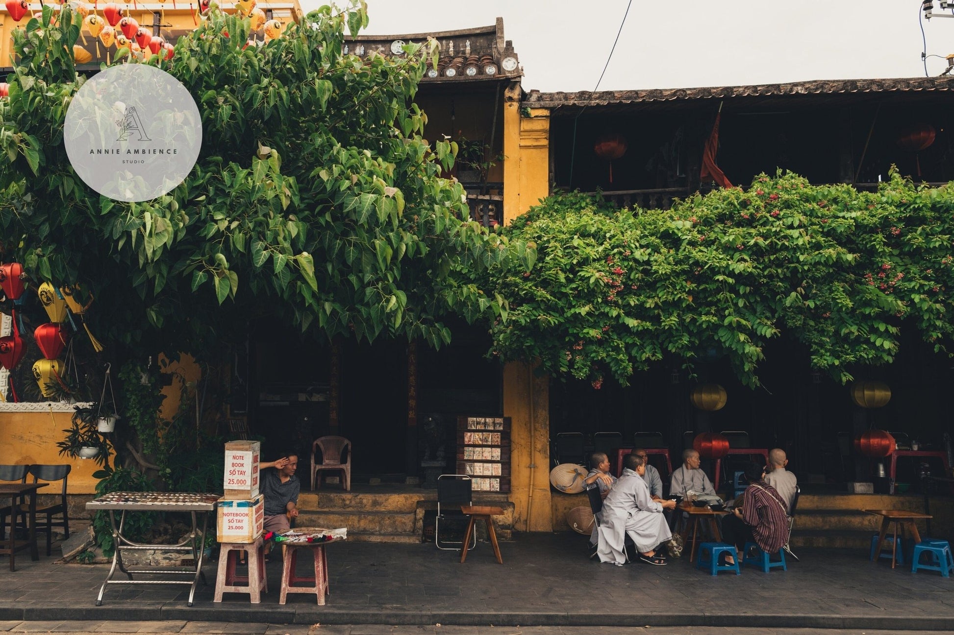 People sit outside a café, embrace Afternoon in Hoi An with lush greenery, dark wooden tables, and yellow walls.