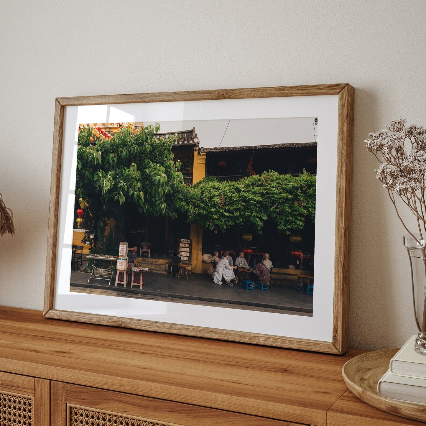 The Afternoon in Hoi An framed photo, depicting trees and people, is displayed on a wooden table near dried flowers.