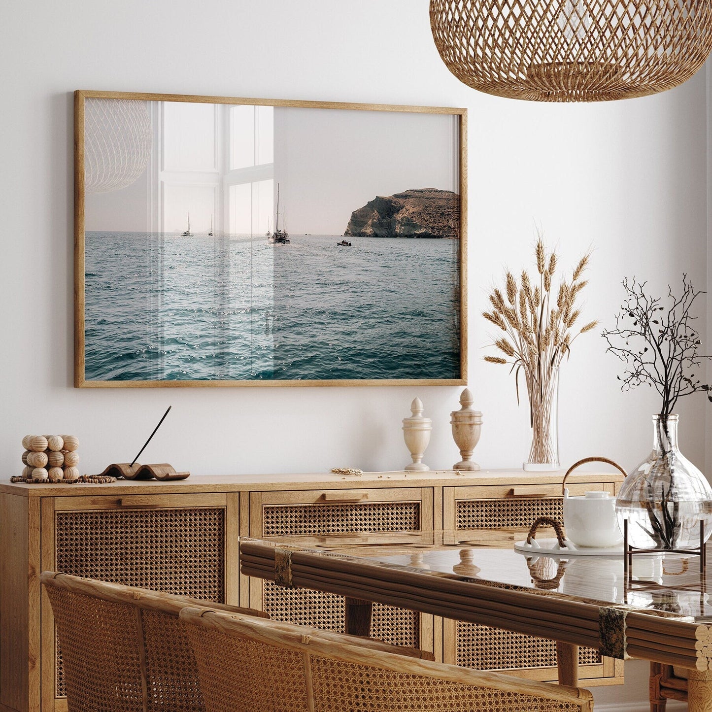 A dining room with a wicker table, rattan sideboard, Afternoon Sail II photo, glass decor, and dried plants in vases.