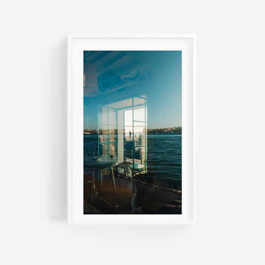 Almada Reflections: Framed photo capturing indoor chairs and seaside views through a window’s reflection.