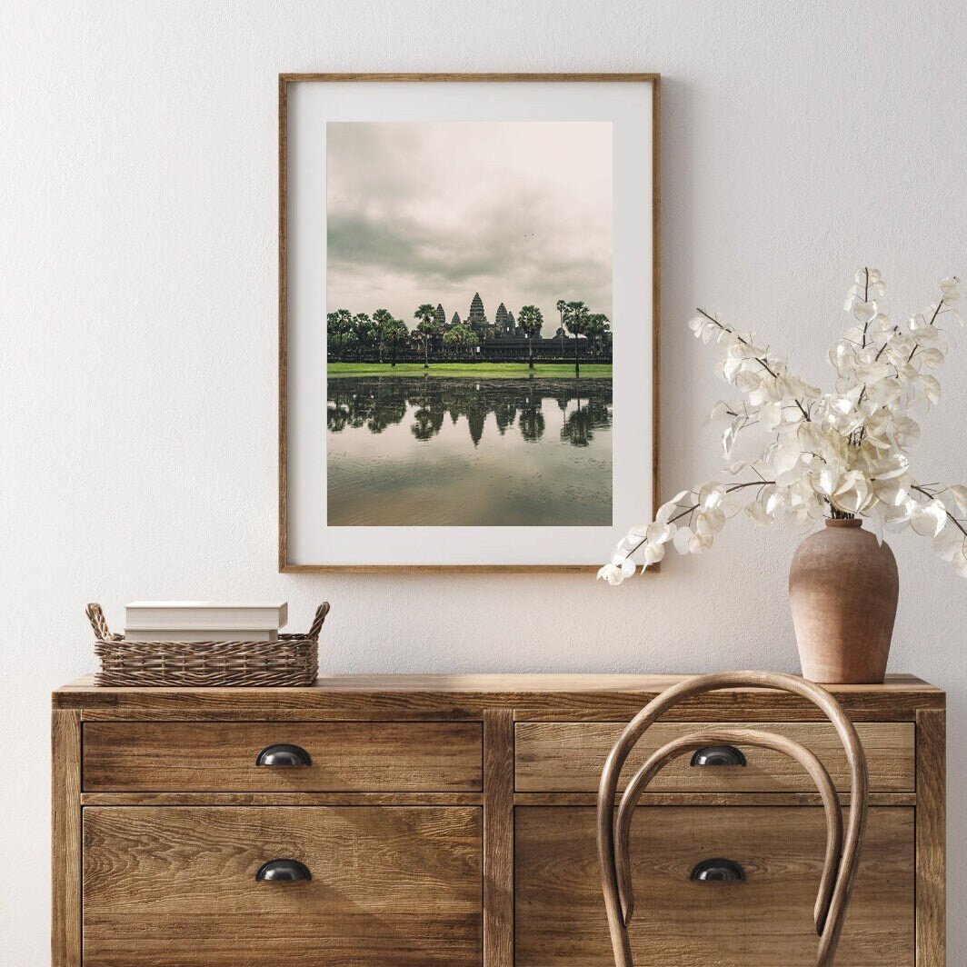 The Angkor Wat framed landscape photo hangs above a wooden dresser with a wicker basket and vase of white flowers.
