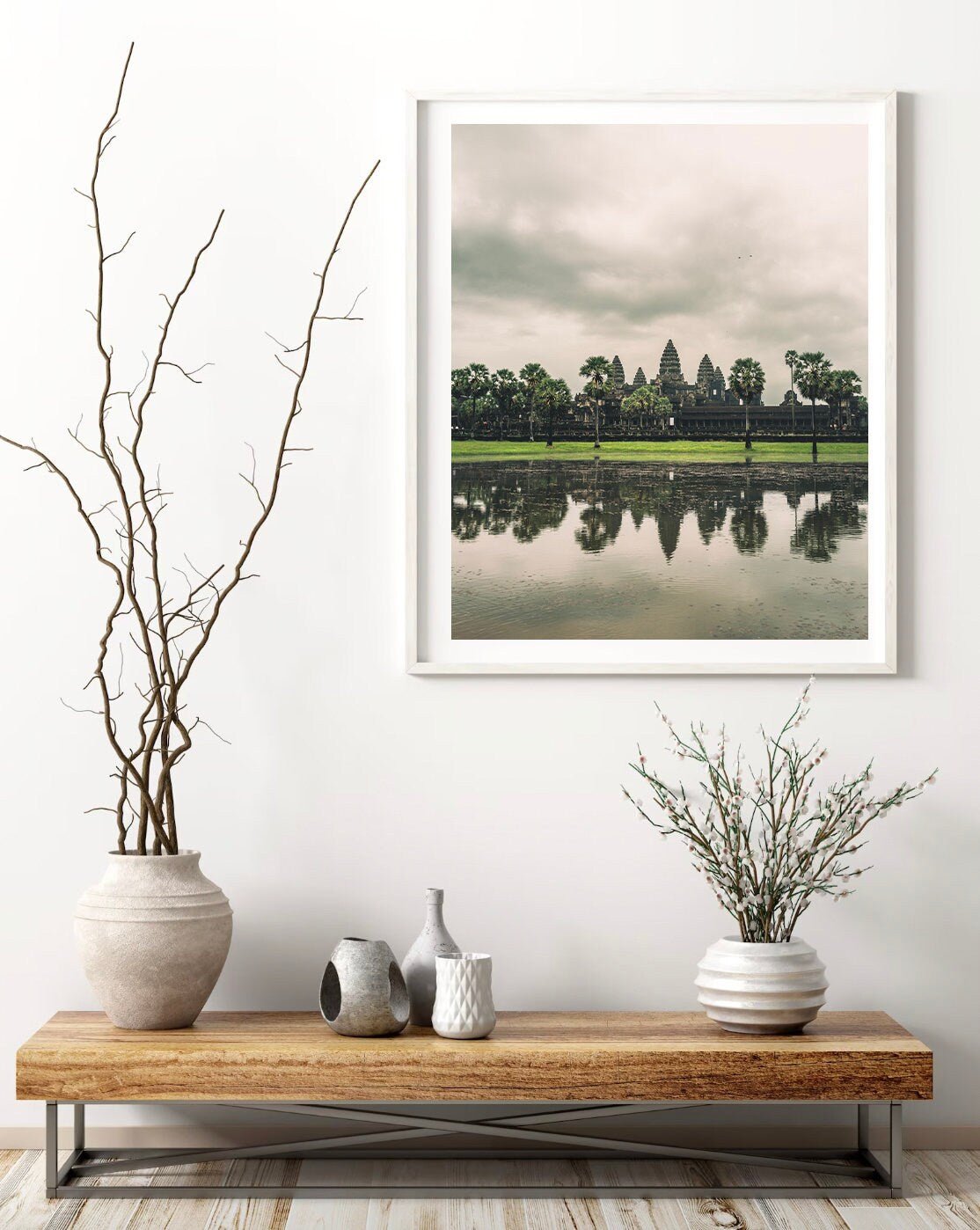 A serene room with Angkor Wat on a wooden bench and a framed landscape photo hung on a white wall.