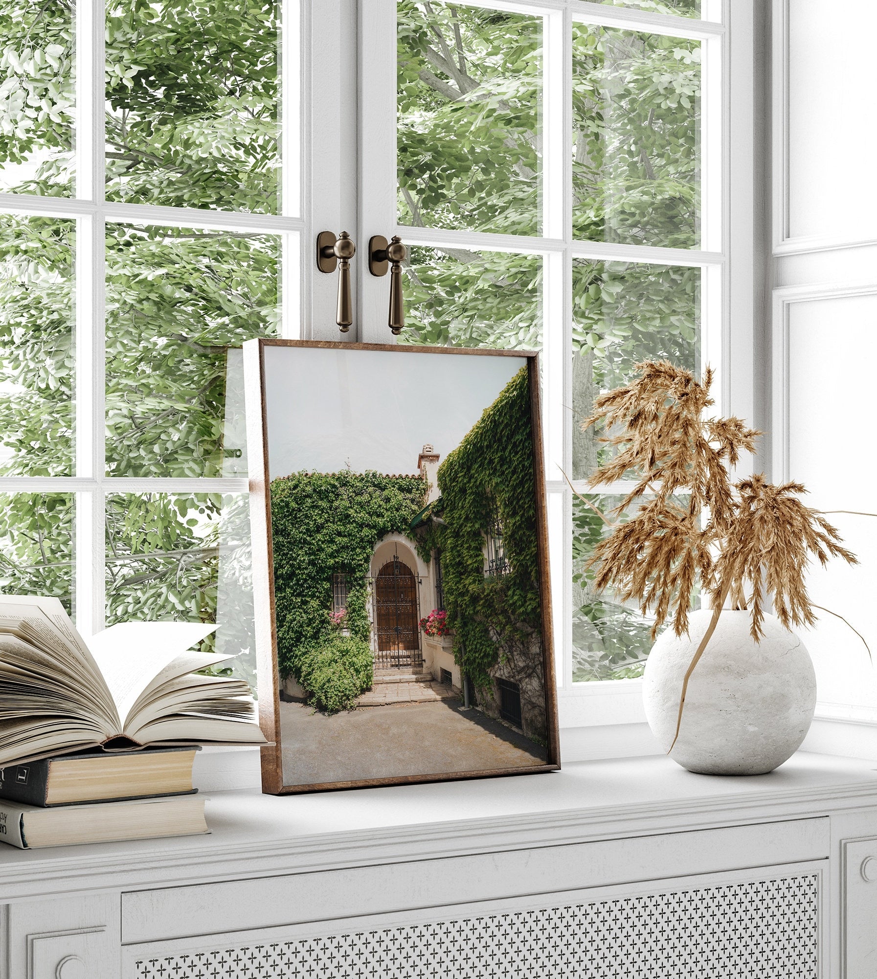 The Basque Oasis picture, framed, sits on a windowsill with books and a vase of dried plants.