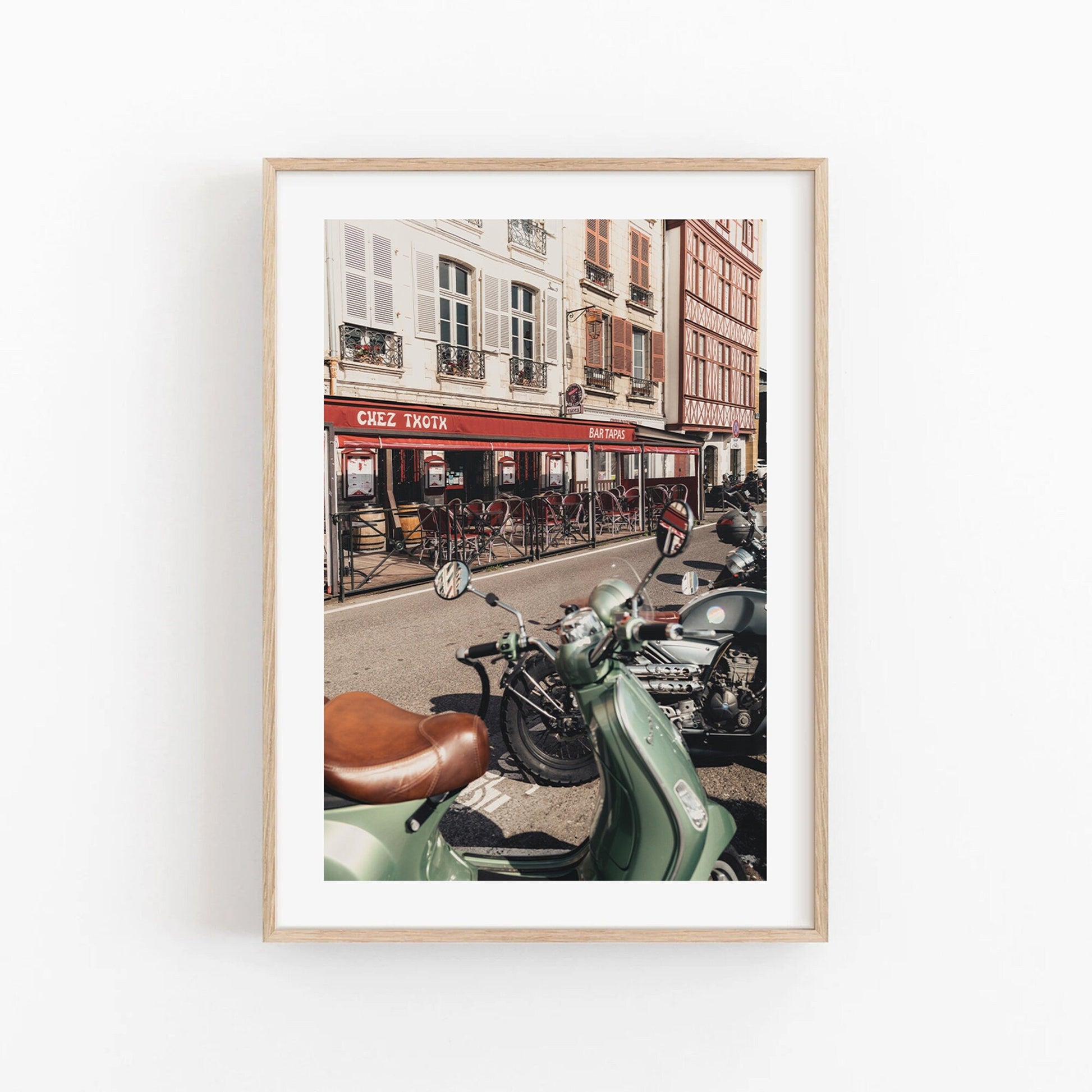 Framed photo Bayonne Vespa: street with parked scooters in front of a café with a red awning and outdoor seating.