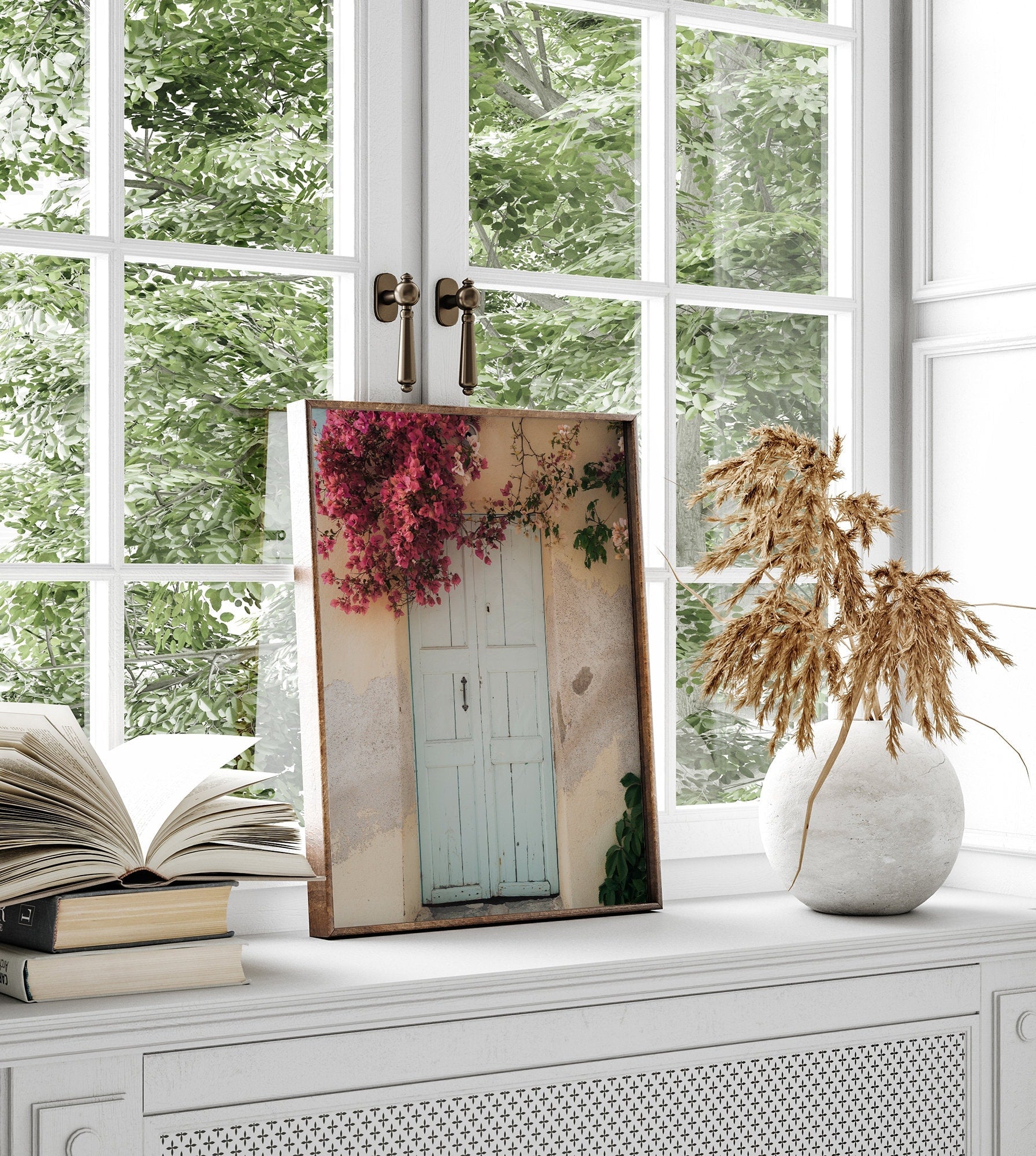 A framed Blue Door Bougainvillea picture sits on a windowsill next to an open book and dry plant.