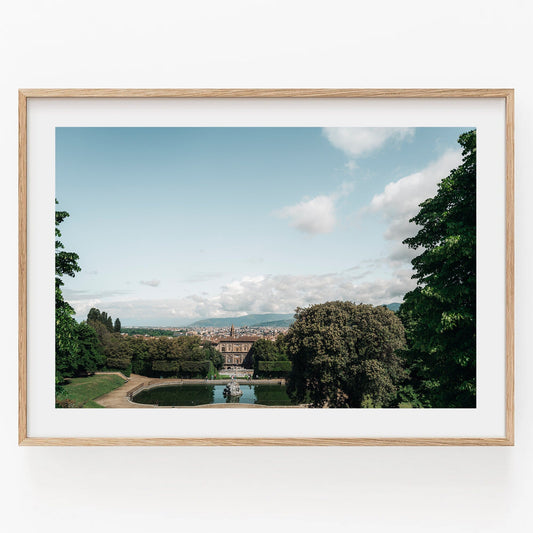 Framed photo of Boboli Gardens with a central fountain and distant cityscape under a partly cloudy sky.