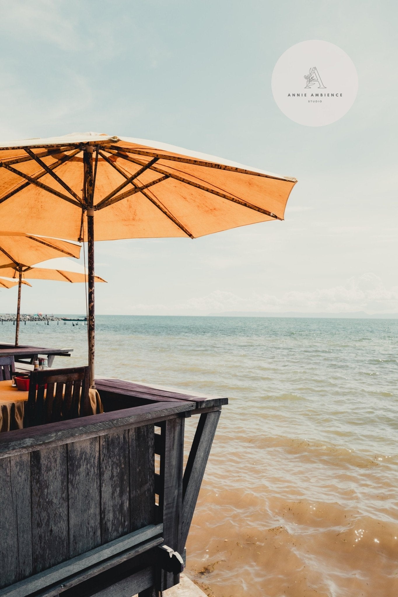The Bright Shores - Set of 3 offers tables under tan parasols overlooking a calm sea on a sunny day.