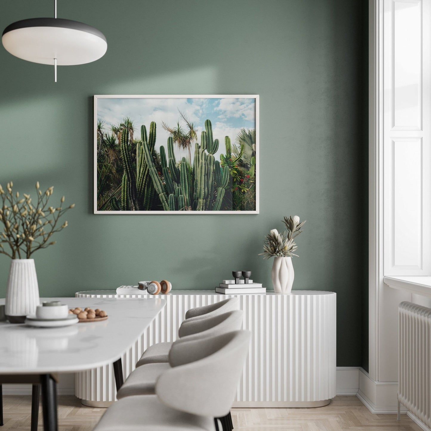A modern dining room with green walls, Cactus Oasis art, a white table, gray chairs, and minimal decor on a white sideboard.