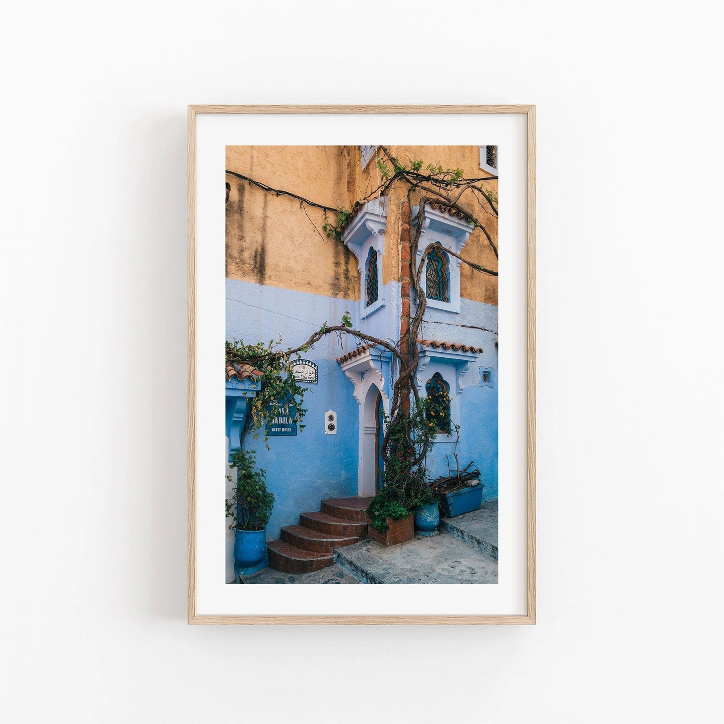 Framed photo titled Chefchaouen II featuring a building with blue, yellow hues and climbing plants.