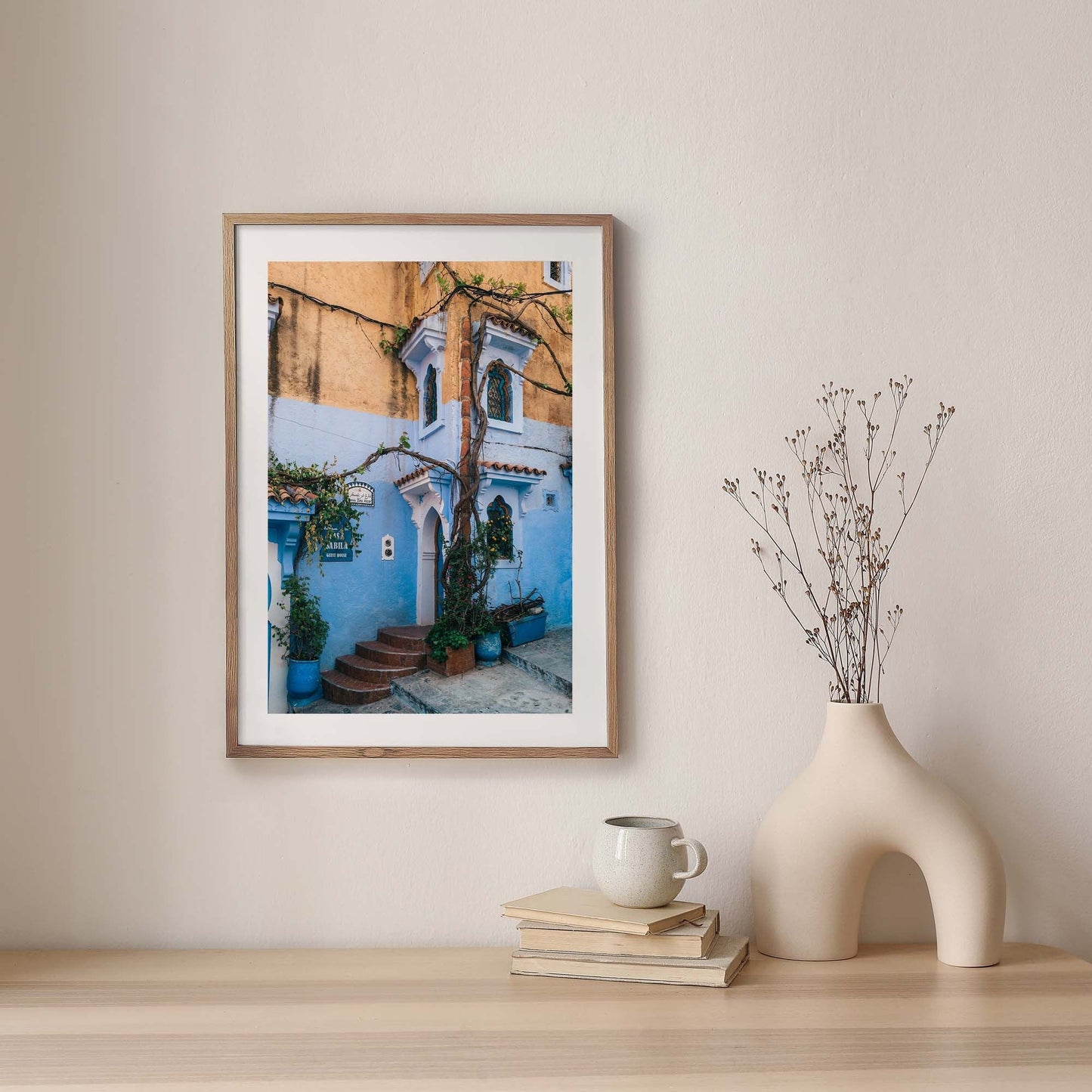 Chefchaouen II photo framed, featuring a vibrant blue and orange building, adorns a wall with vases, mugs, and books.