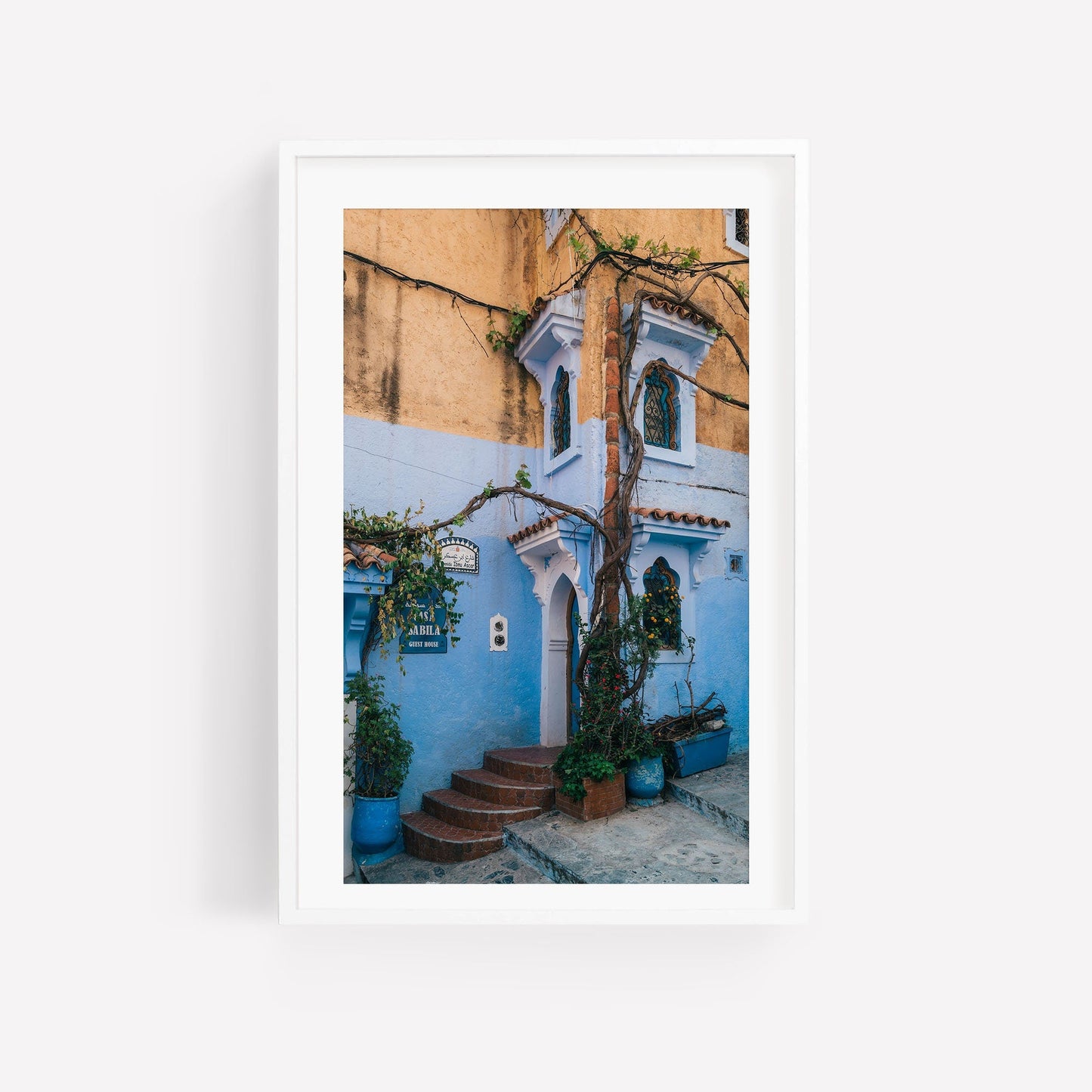 Framed photo titled Chefchaouen II featuring a charming blue house with a tree and arched doorways against a light blue wall.