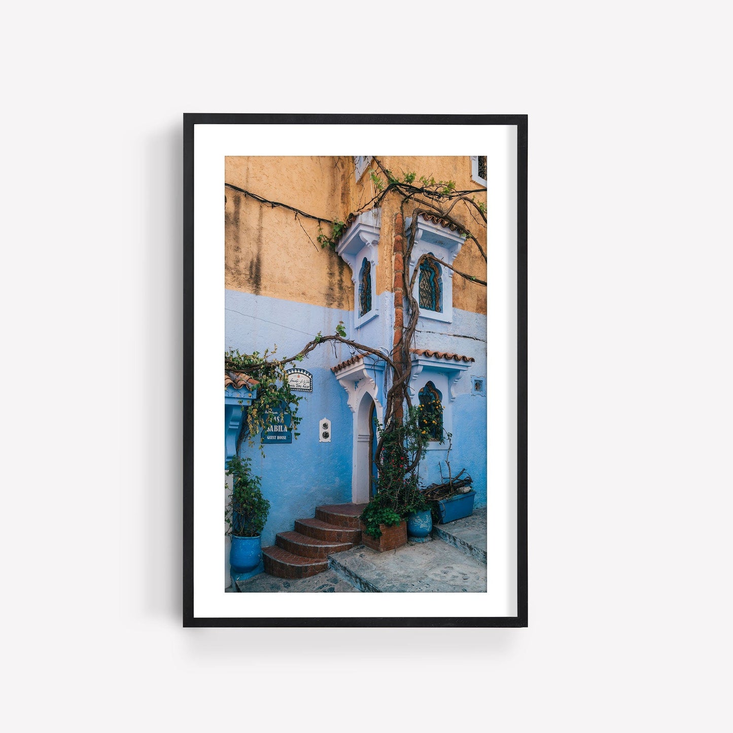 Framed photo titled Chefchaouen II featuring a blue and yellow building with plants, arched windows, and a small staircase.
