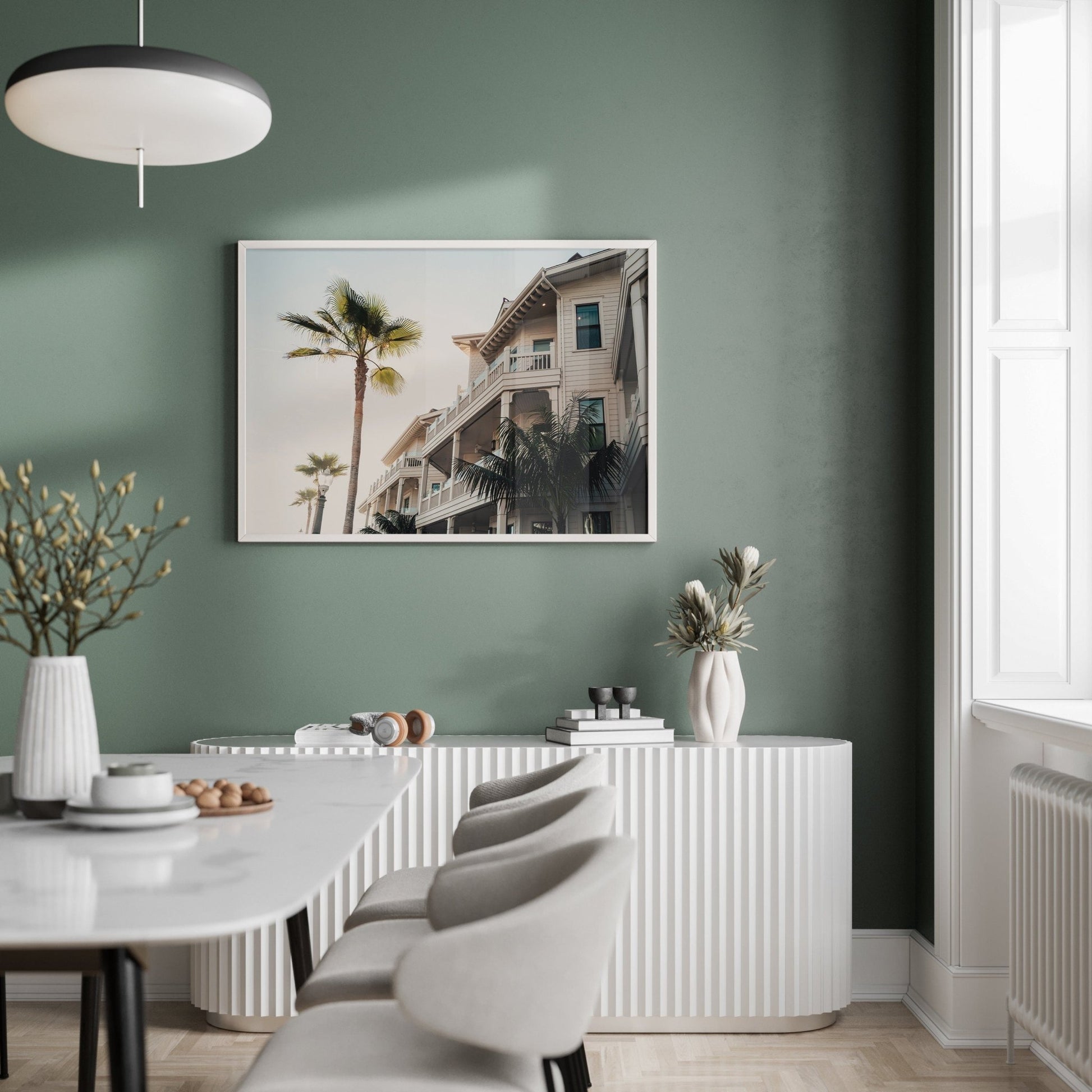 Modern dining room featuring a green wall, white furniture, and the Coastal Abode framed photo of palm trees and houses.