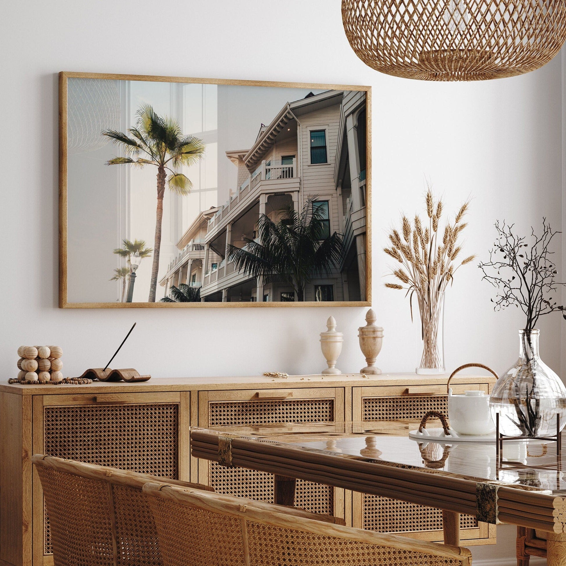 Dining room with Coastal Abode, wicker furniture, a large framed photo of a house with palm trees, and vases on a buffet.