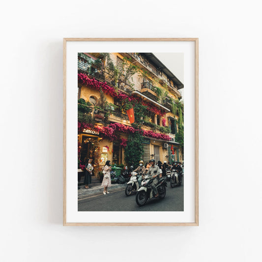 Colors of Hanoi framed photo features a vibrant street scene with colorful flowers and parked scooters by a bustling building.