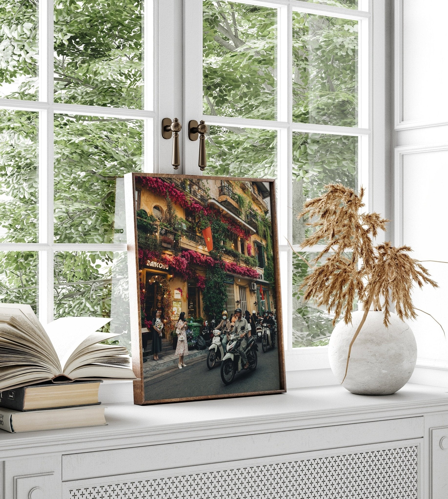 Colors of Hanoi framed photo on window ledge with book and vase, greenery outside.