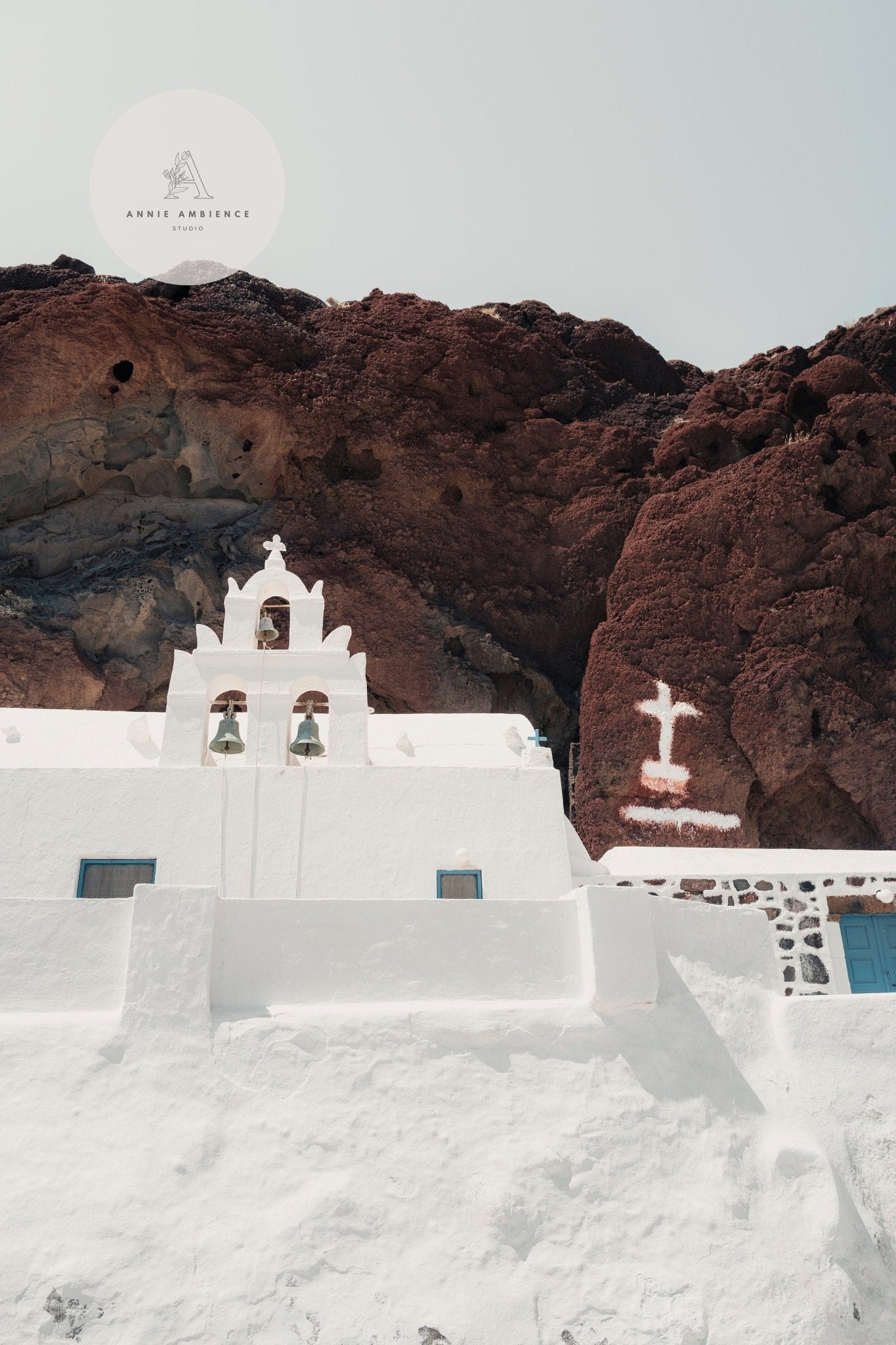 White church with bell tower and blue doors against cliffs, under clear skies—Colors of Santorini.