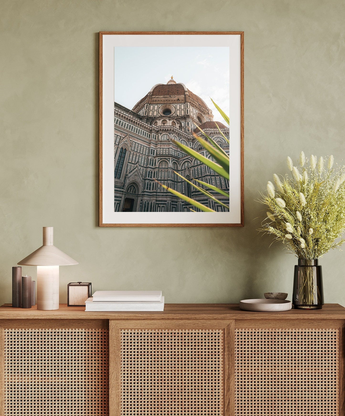 Framed Duomo photo on green wall above a wicker cabinet with decor and flower vase.