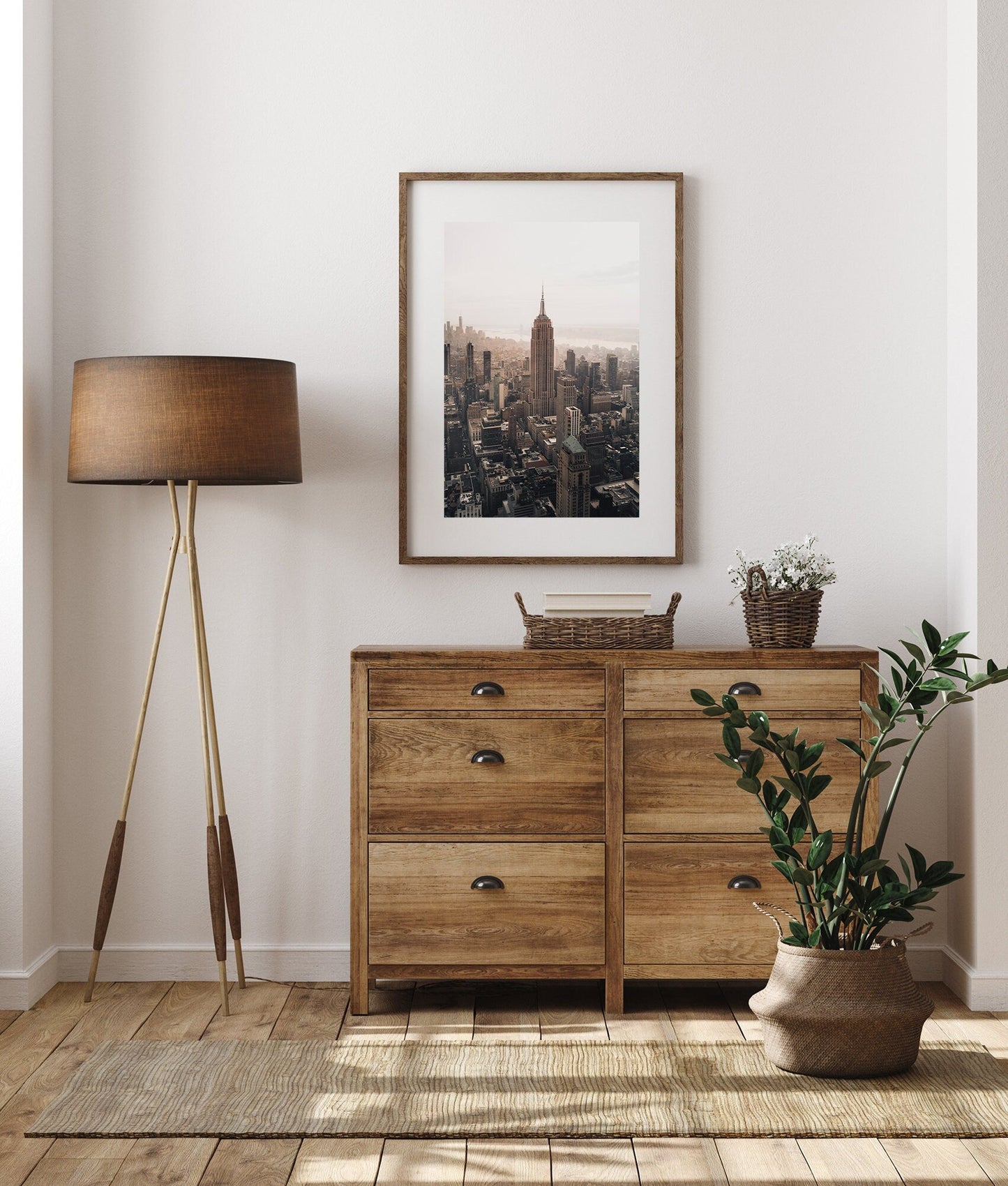 A cozy room with a wooden dresser, standing lamp, framed Empire State Building, potted plant on wooden flooring.