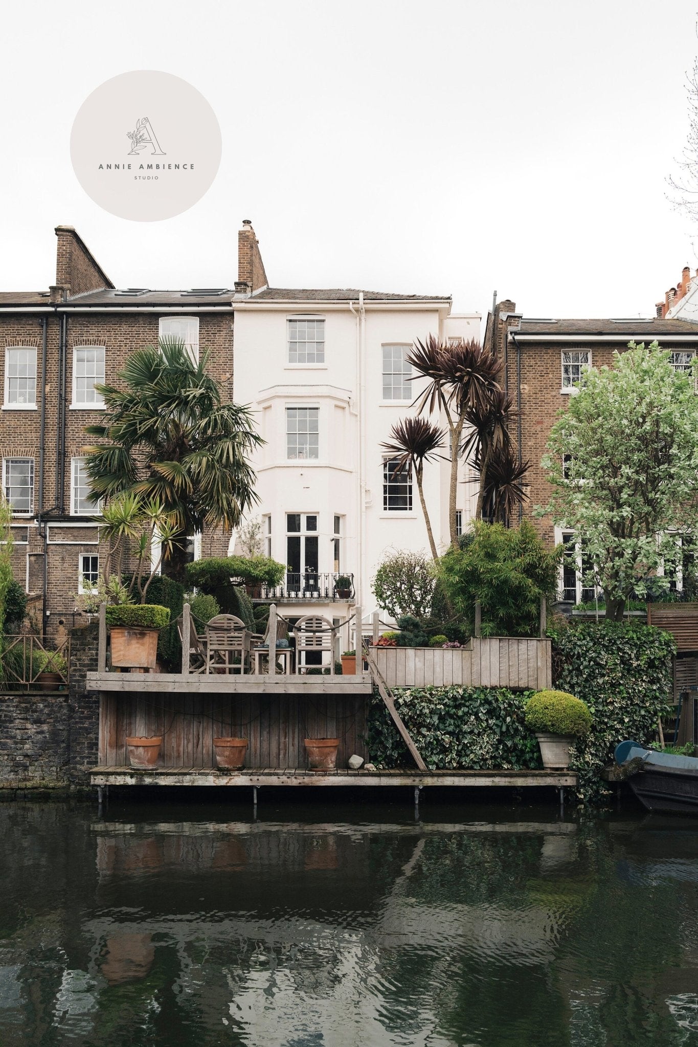 European Charm - Set of 3: A serene riverside with a brick house, lush greenery, and seating facing the water.