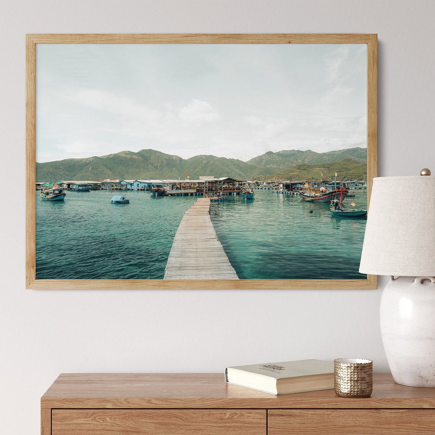 Fishing Village I framed photo, wooden pier to boats on a lake, displayed above a table with lamp and book.