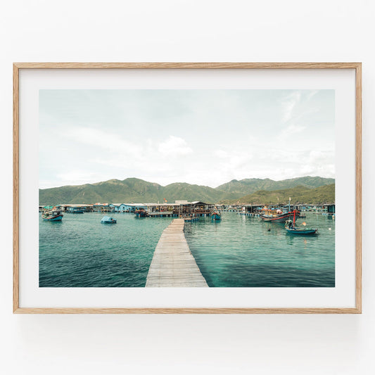 Framed Fishing Village I photo: wooden dock over calm water, small boats, and mountain scenery.