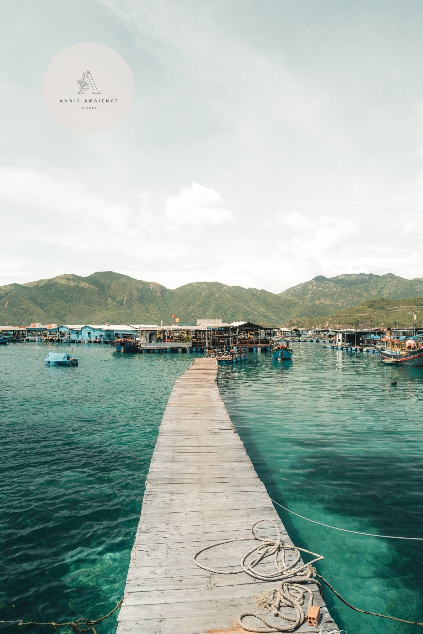 Fishing Village II: A wooden pier leads over turquoise water to floating structures with green mountains behind.