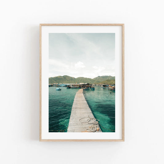 Framed photo Fishing Village II shows a wooden pier stretching over calm waters towards mountains and a clear sky.