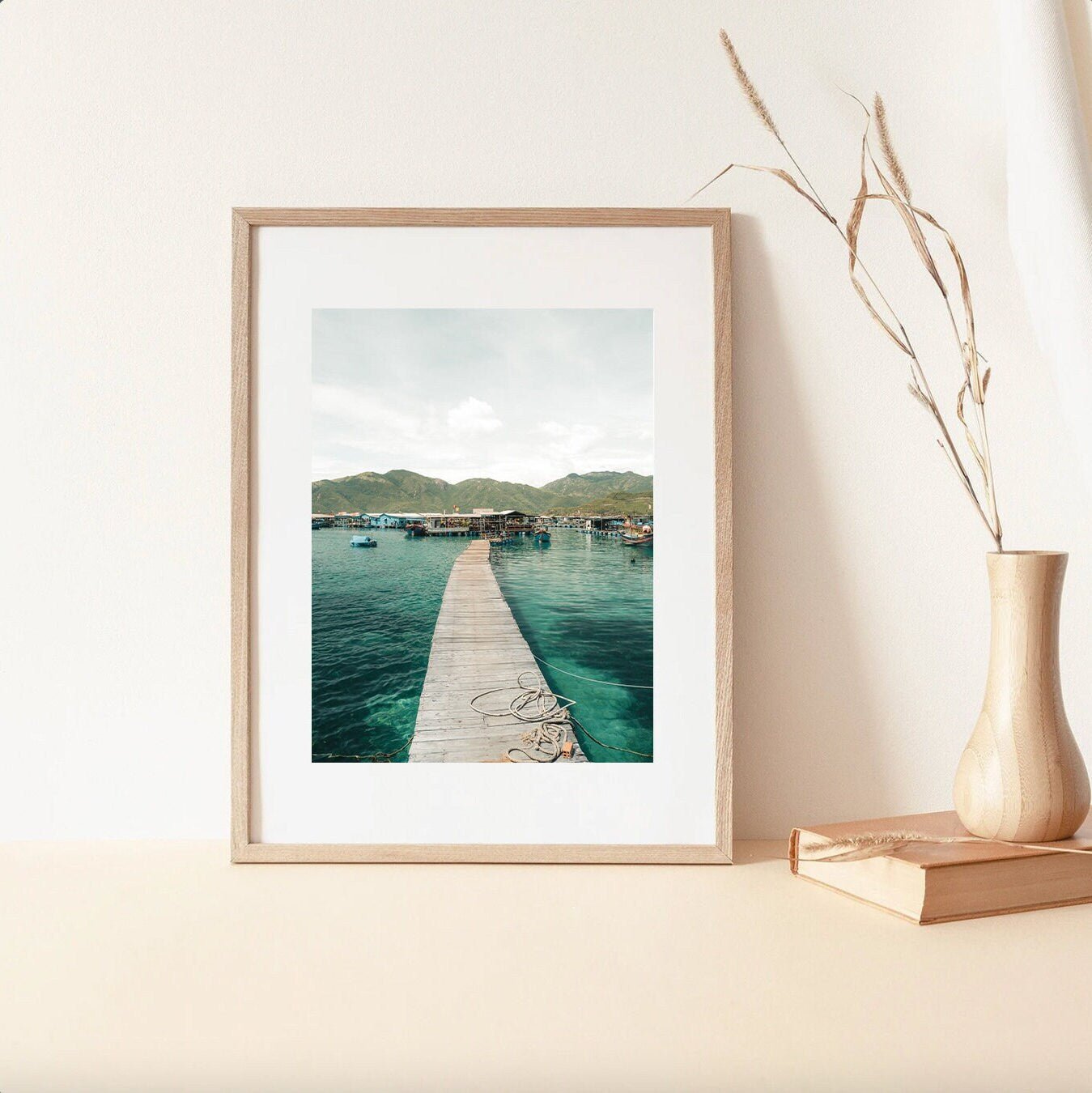 Fishing Village II framed photo on a light table next to a vase with dry grass.