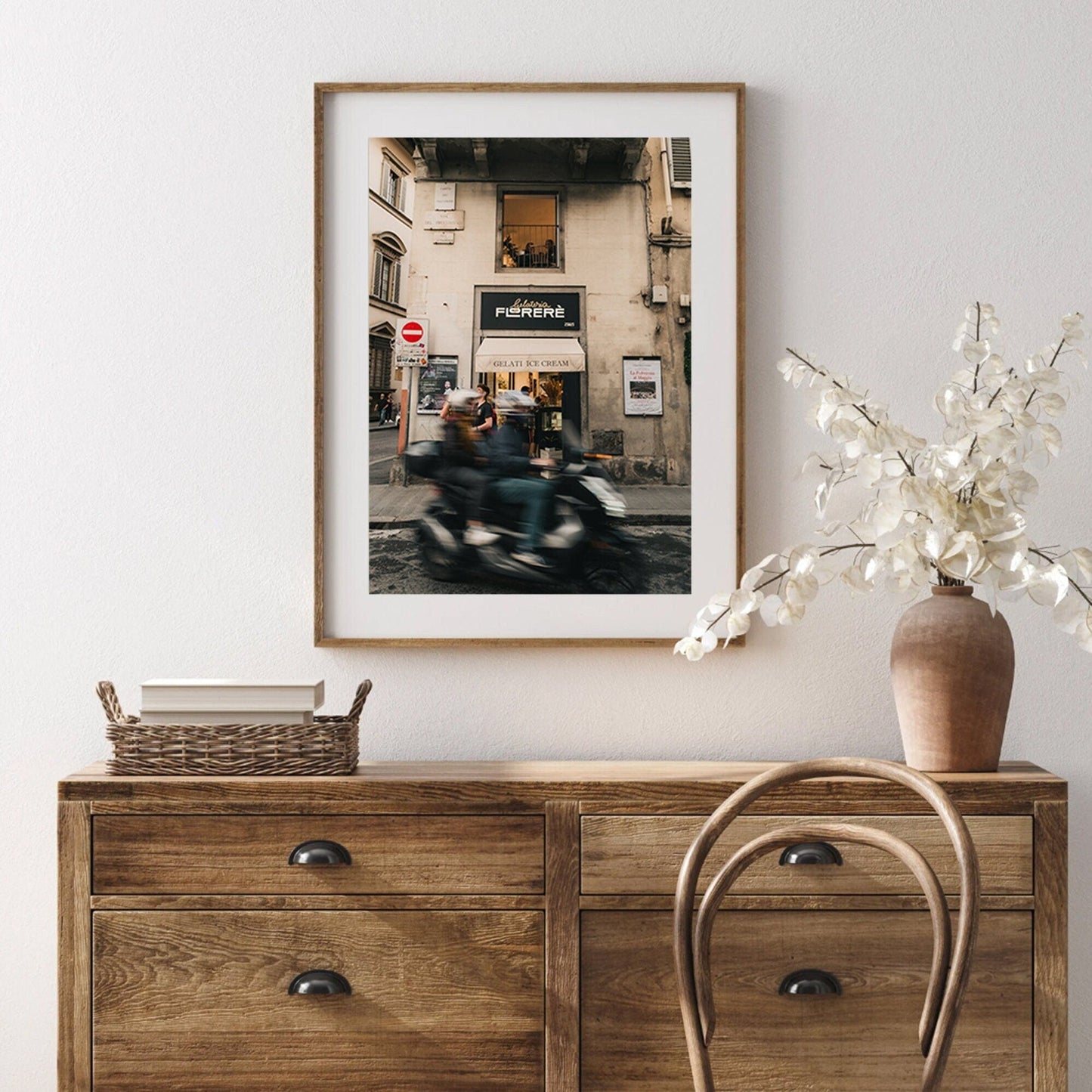A framed street photo hangs above a wooden dresser with the Florence Motorcycle, a wicker basket, and white flowers.