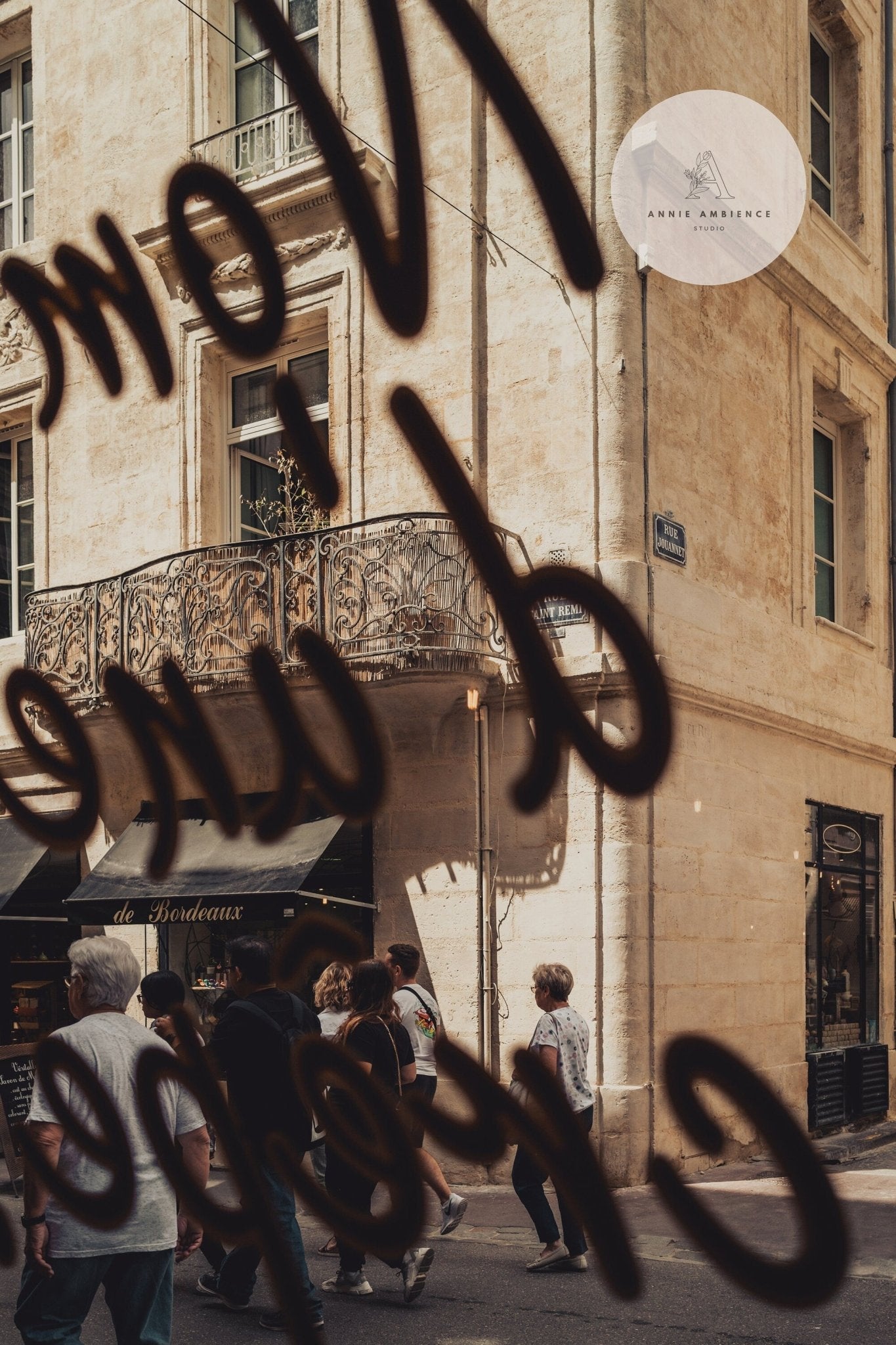 People stroll past a beige building with a wrought iron balcony, featuring French Charm text on the glass in front.
