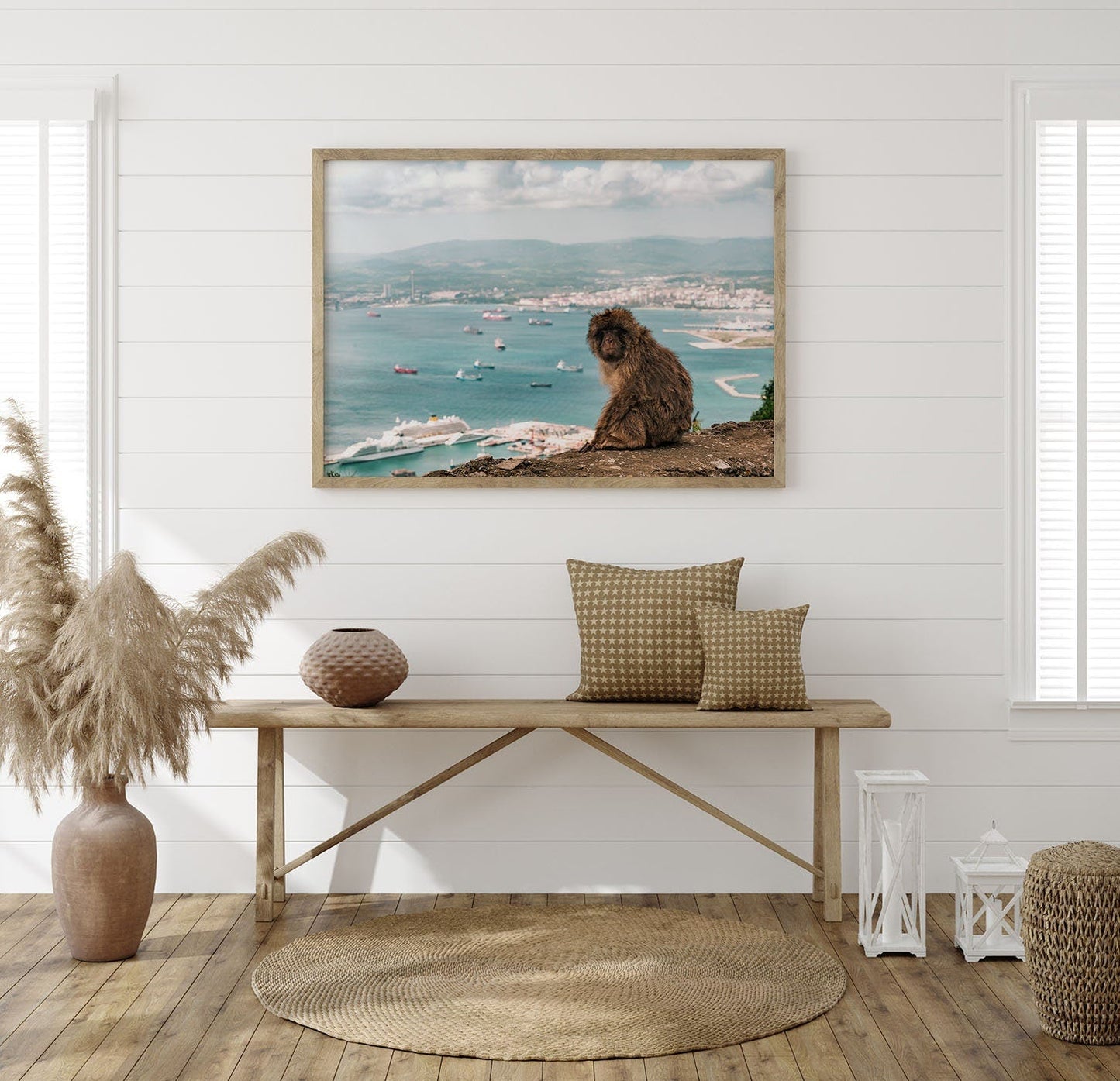 A cozy room with a rattan bench, pillows, vase, and Gibraltar Monkey wall art overlooking a coastal city and sea.