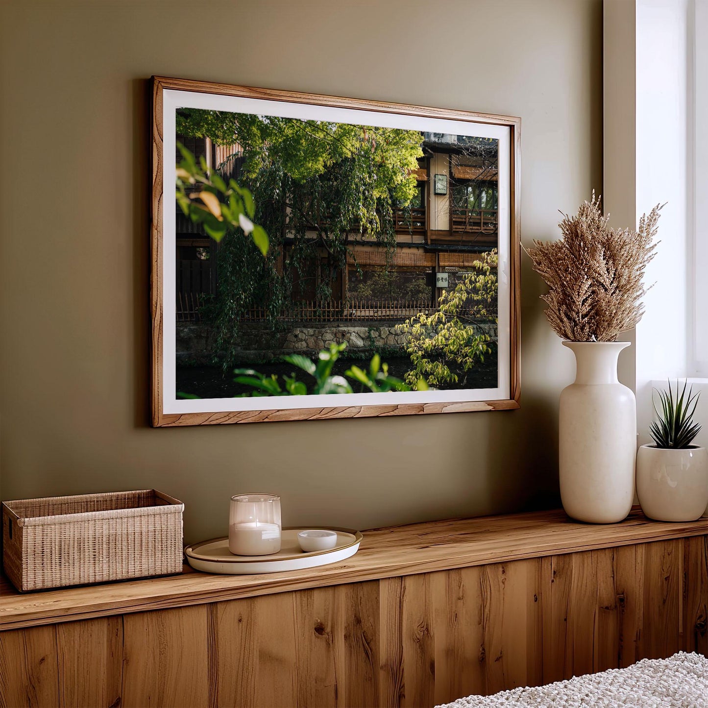 The Gion Greenery framed picture showcases a house, set on a green wall with plants and a candle adorning the wooden sideboard.