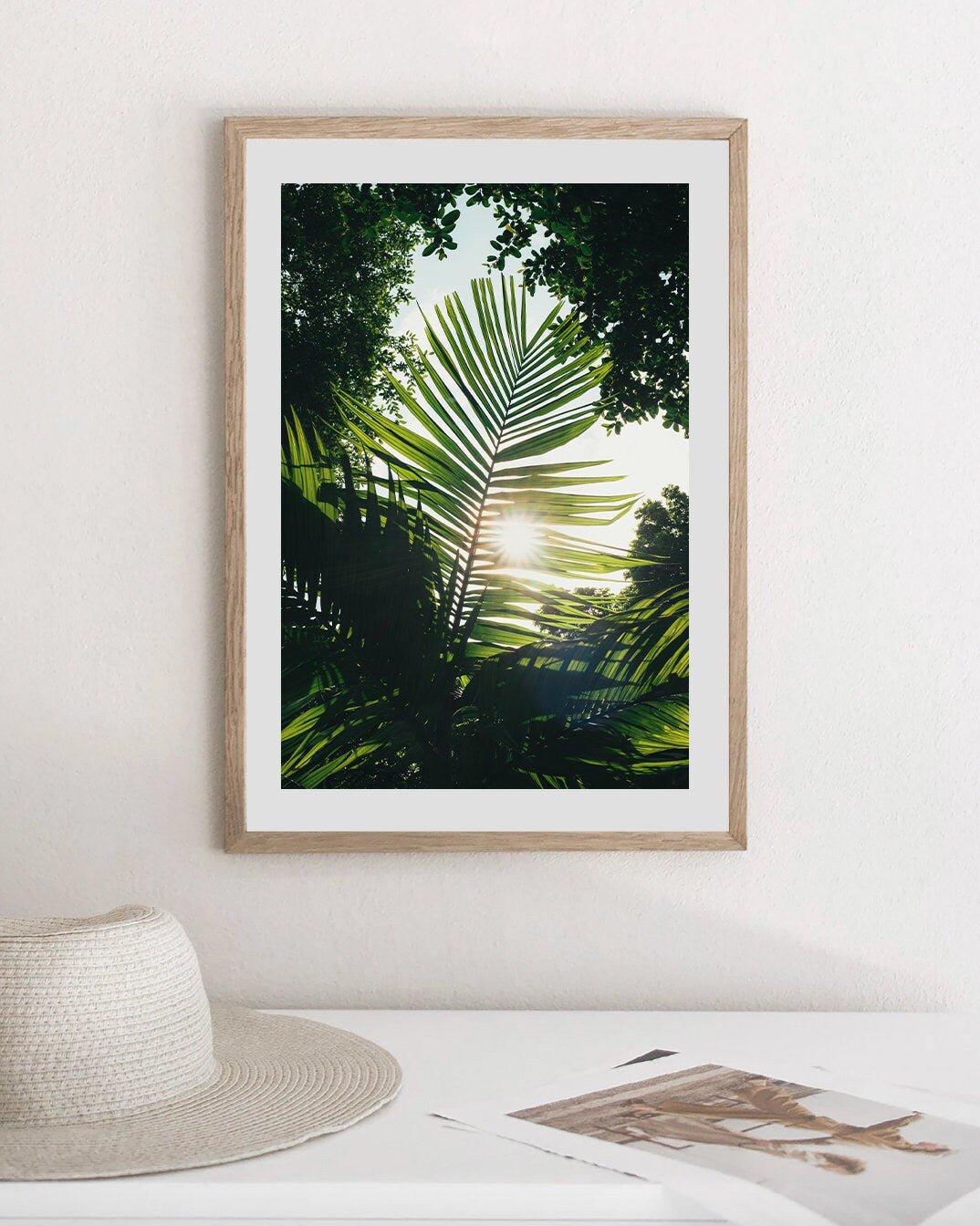 The Glimpse of Hope picture of palm leaves hangs above a table with a hat and papers on a white wall.