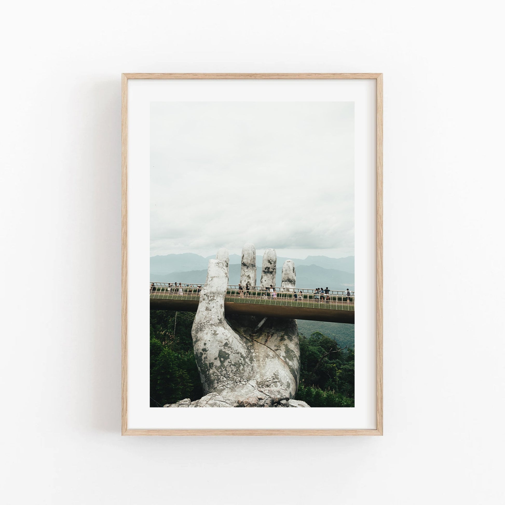 Framed photo titled Golden Hand Bridge I depicts a stone hand holding a pedestrian bridge with tourists against clouds and mountains.