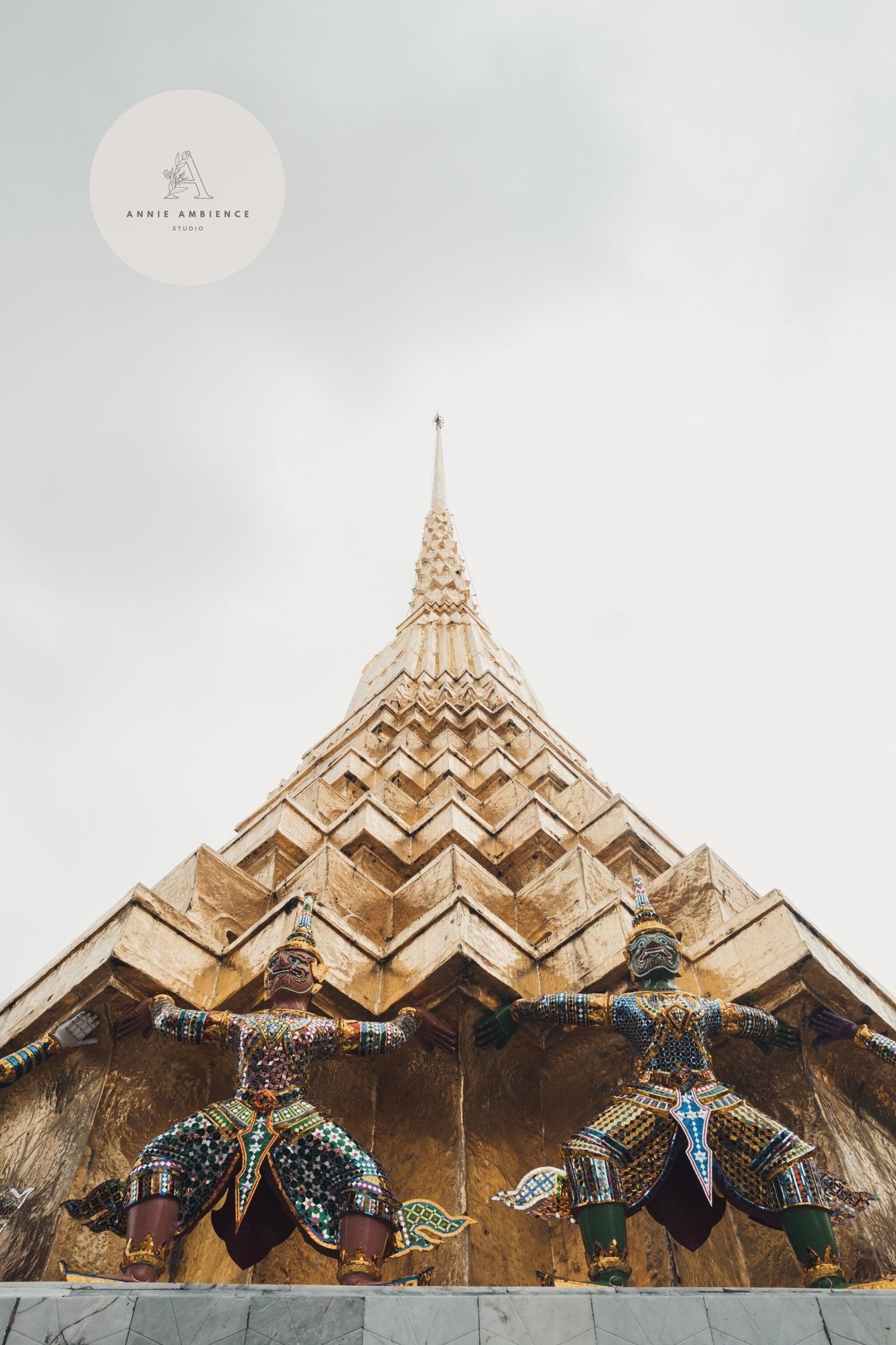 View up at the Grand Palace Is golden spire with two ornate statues at the base against a cloudy sky.