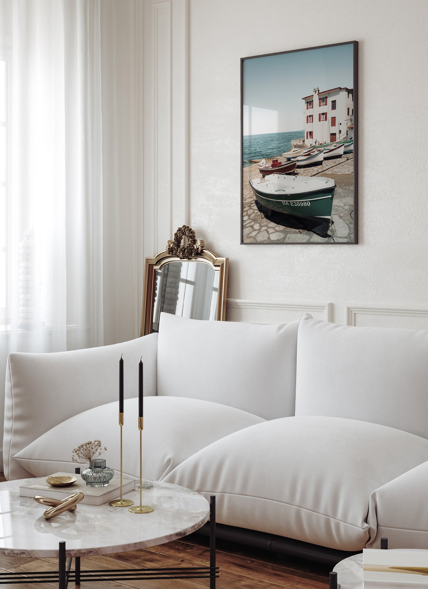 A bright living room with a white sofa, marble table, and Guethary wall art of boats by a seaside building.