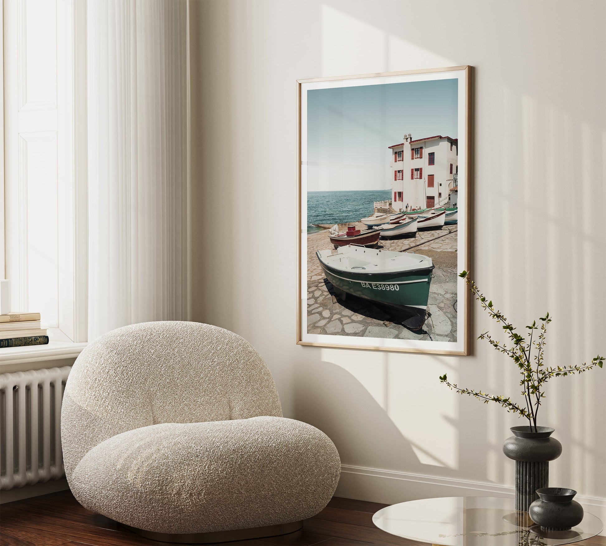 The Guethary cream chair sits by a round table, vase, and framed seaside boat artwork on the wall.
