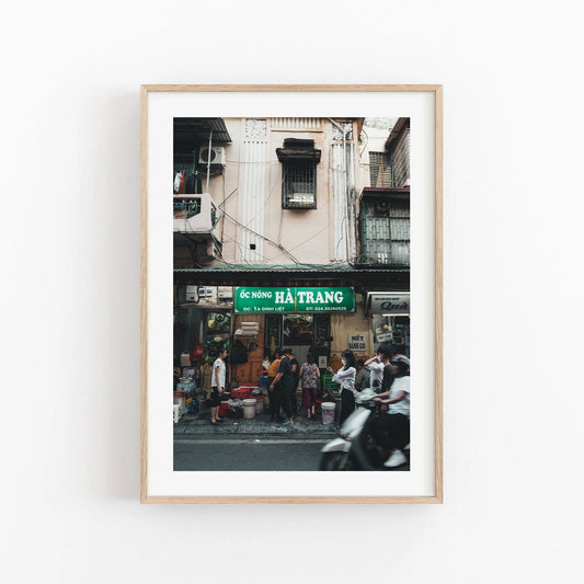 Framed Bustling Hanoi print: a street scene with people and motorbikes outside a shop in Vietnam.