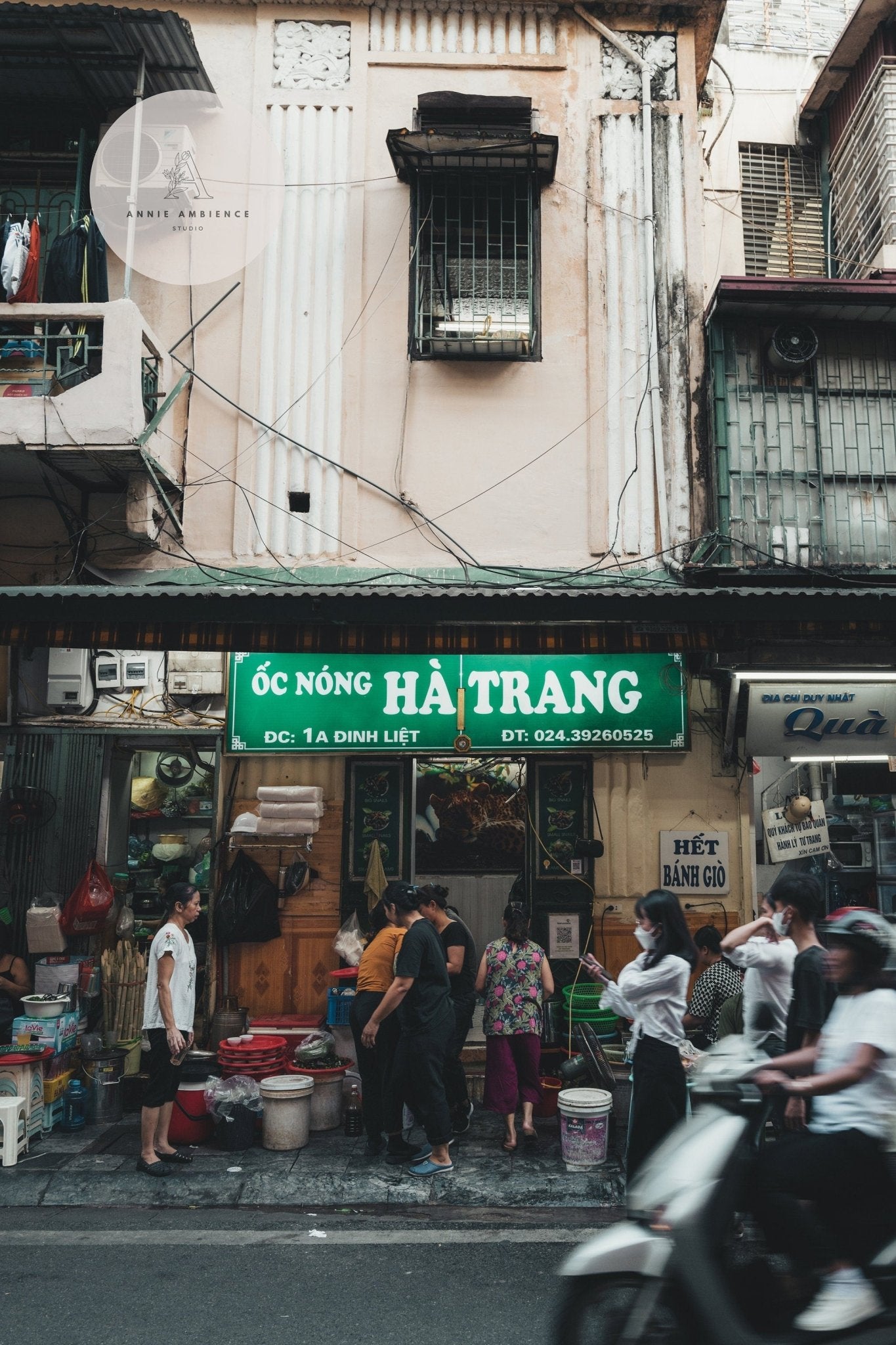 Street scene of a small, bustling Hanoi seafood shop in a vibrant alley with pedestrians and a motorbike passing by.