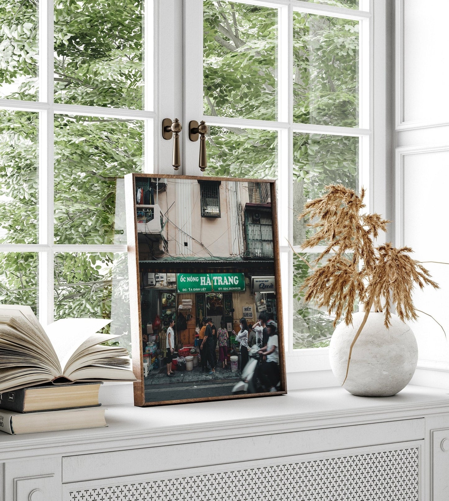 Bustling Hanoi photo framed on a windowsill with books and dried plants in a vase.