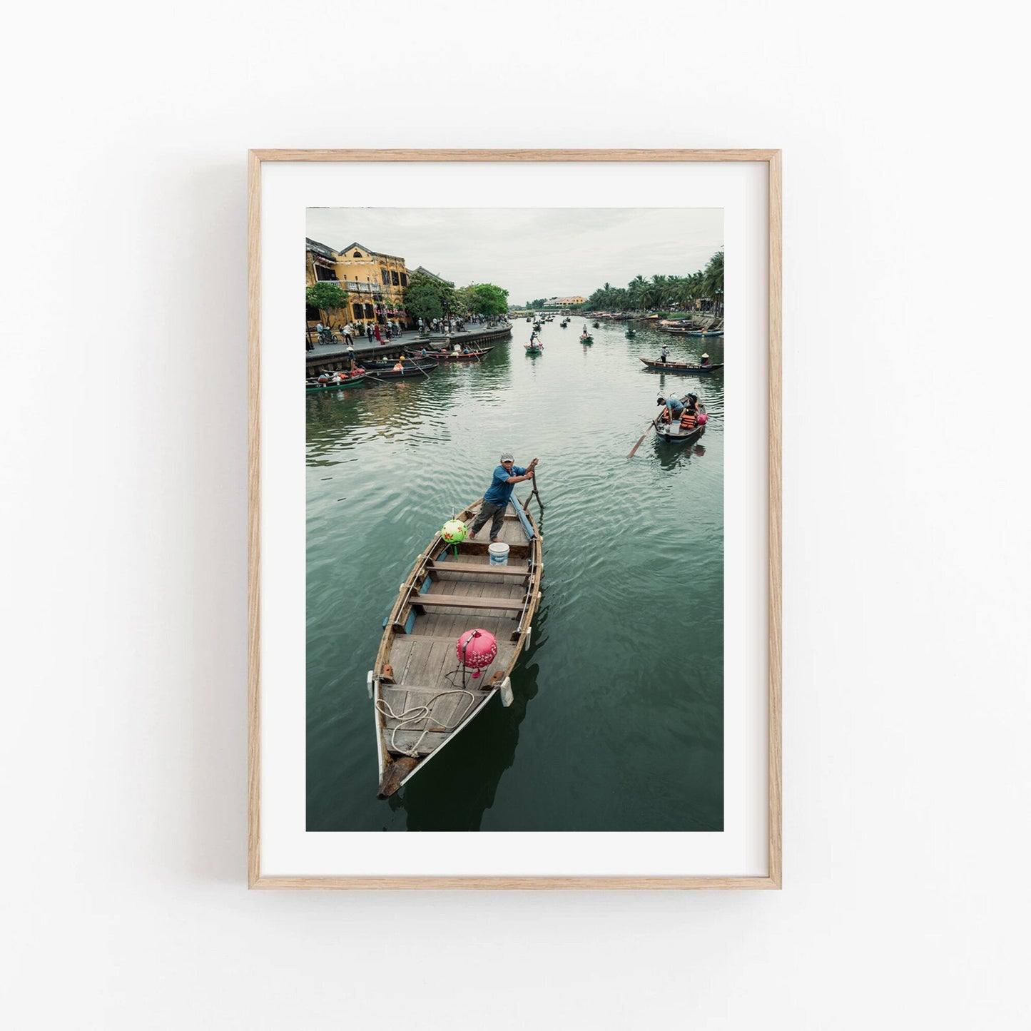Framed photo titled Hoi An Row of a man rowing a wooden boat, with other boats and buildings in the background.