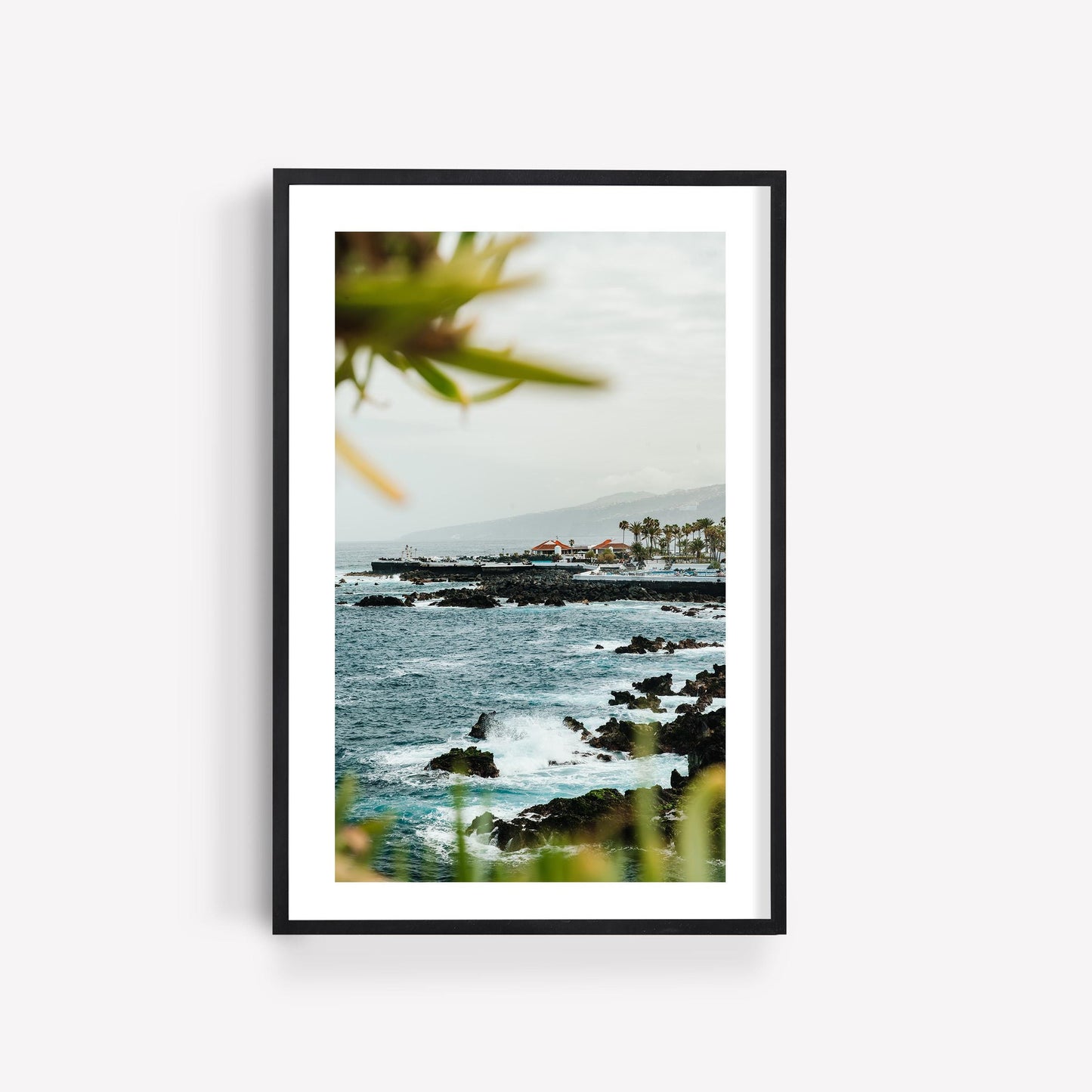 Coastal Glimpse: Framed photo of a rocky coast with waves, distant buildings, and foreground greenery.