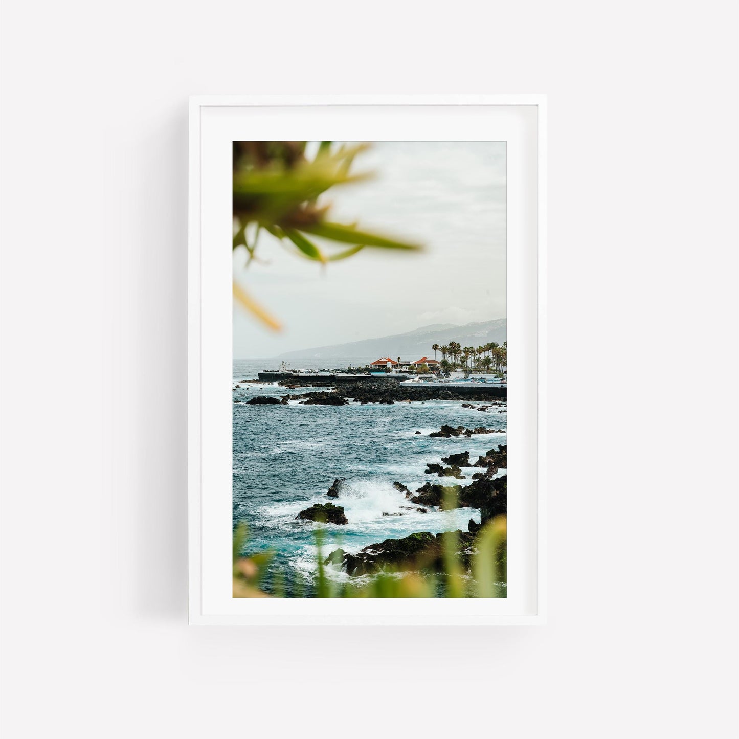 Coastal Glimpse: Framed photo of a rocky coastline with waves, distant buildings, and foreground greenery.