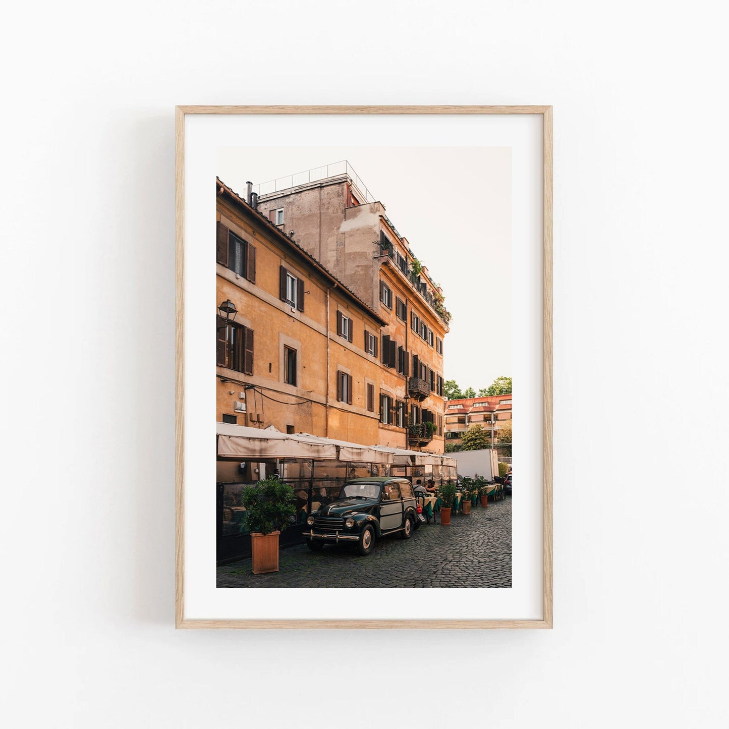 Framed photo of an Italian street featuring a vintage car, buildings, and café tables. Product: Italian Car.