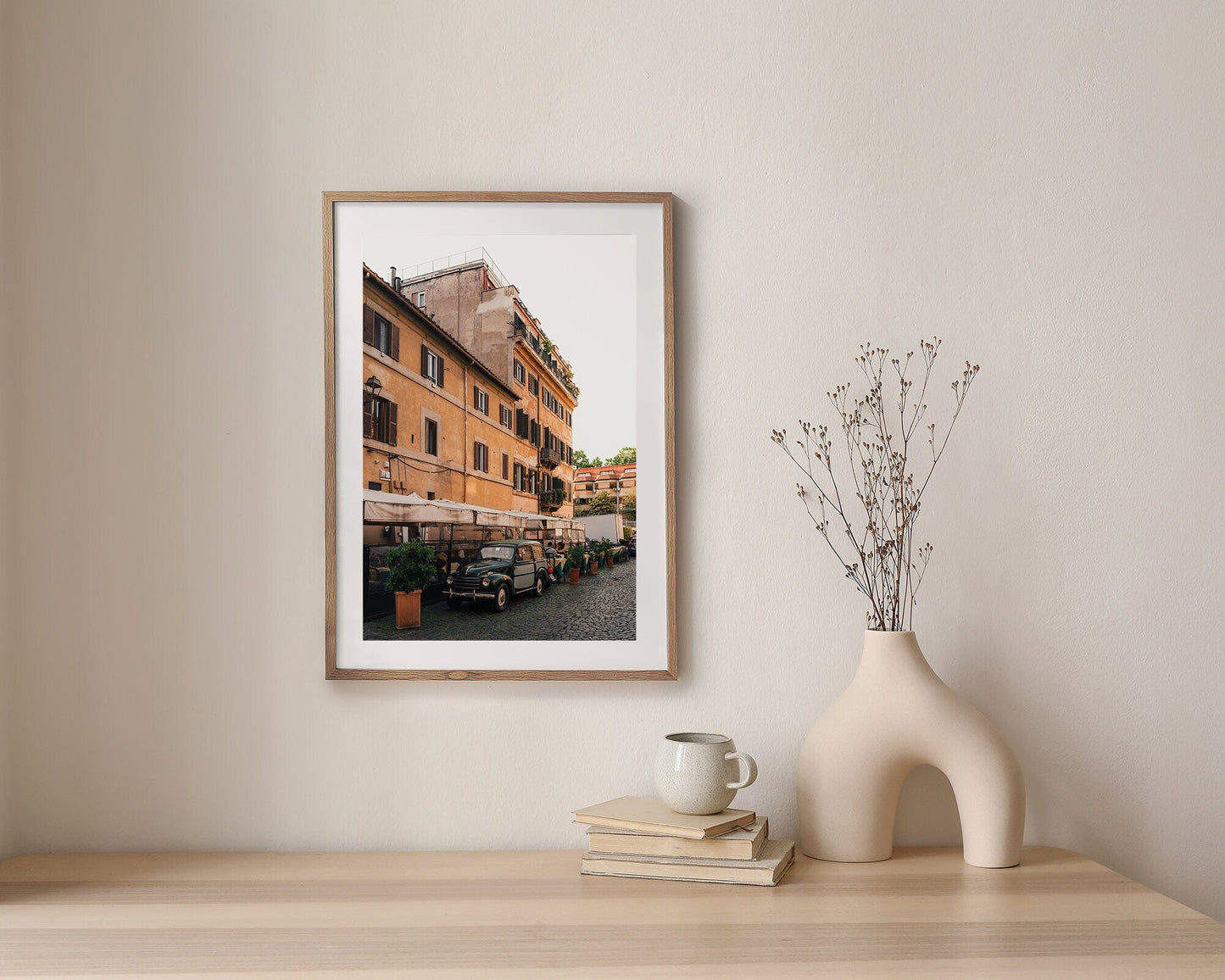 Italian Car photo framed on a wall above a wooden table with a vase, dried plant, books, and a mug.