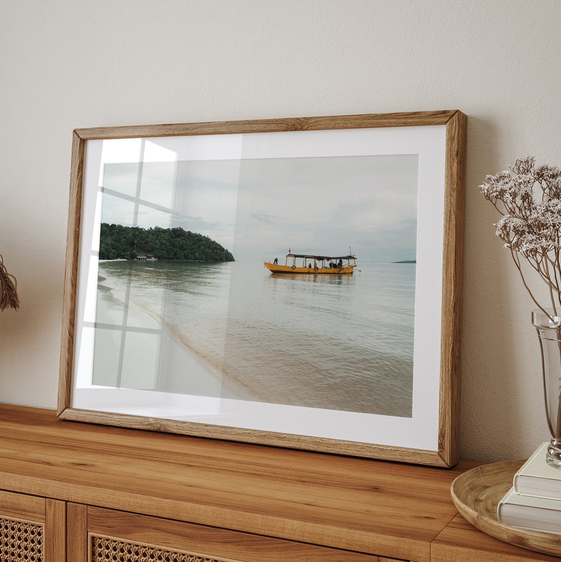 Framed Koh Rong photo of a yellow boat on calm waters, placed on a sideboard with dried flowers in a vase.